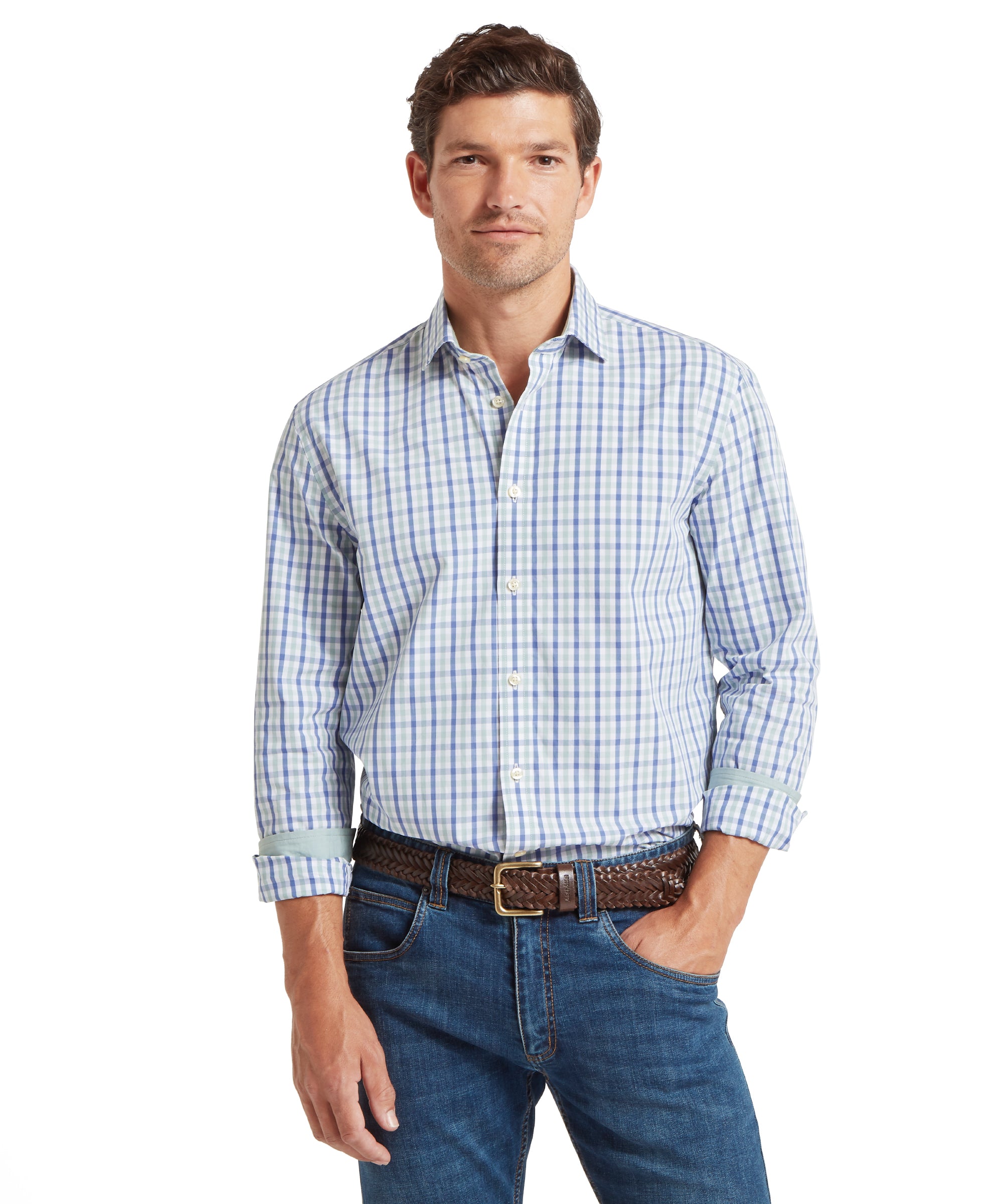 Man wearing a long-sleeved, button-down shirt with a check pattern in shades of blue, green, and white. The shirt is tucked into blue jeans and secured with a brown woven leather belt. The sleeves are rolled up to the forearms. He is standing with one hand in his pocket and the other arm relaxed. The outfit is casual, suitable for everyday or smart-casual occasions.