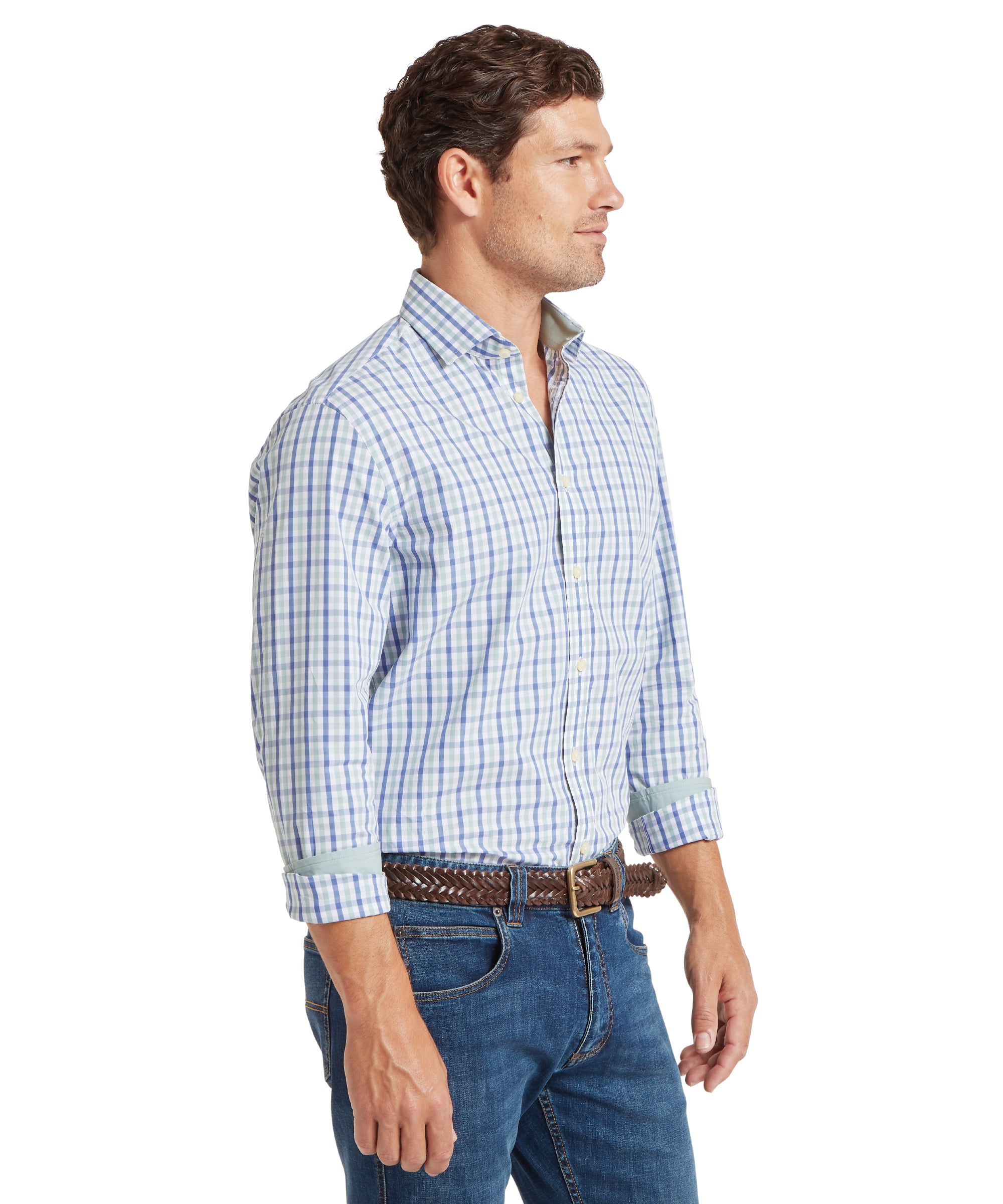 Man standing in profile view, facing right, wearing a long-sleeved, button-up shirt with a check pattern in light blue, green, and white. The shirt is tucked into blue jeans and secured with a brown woven leather belt. The sleeves are rolled up to the forearms. He has short, wavy brown hair. The outfit is casual, suitable for everyday or smart-casual occasions.