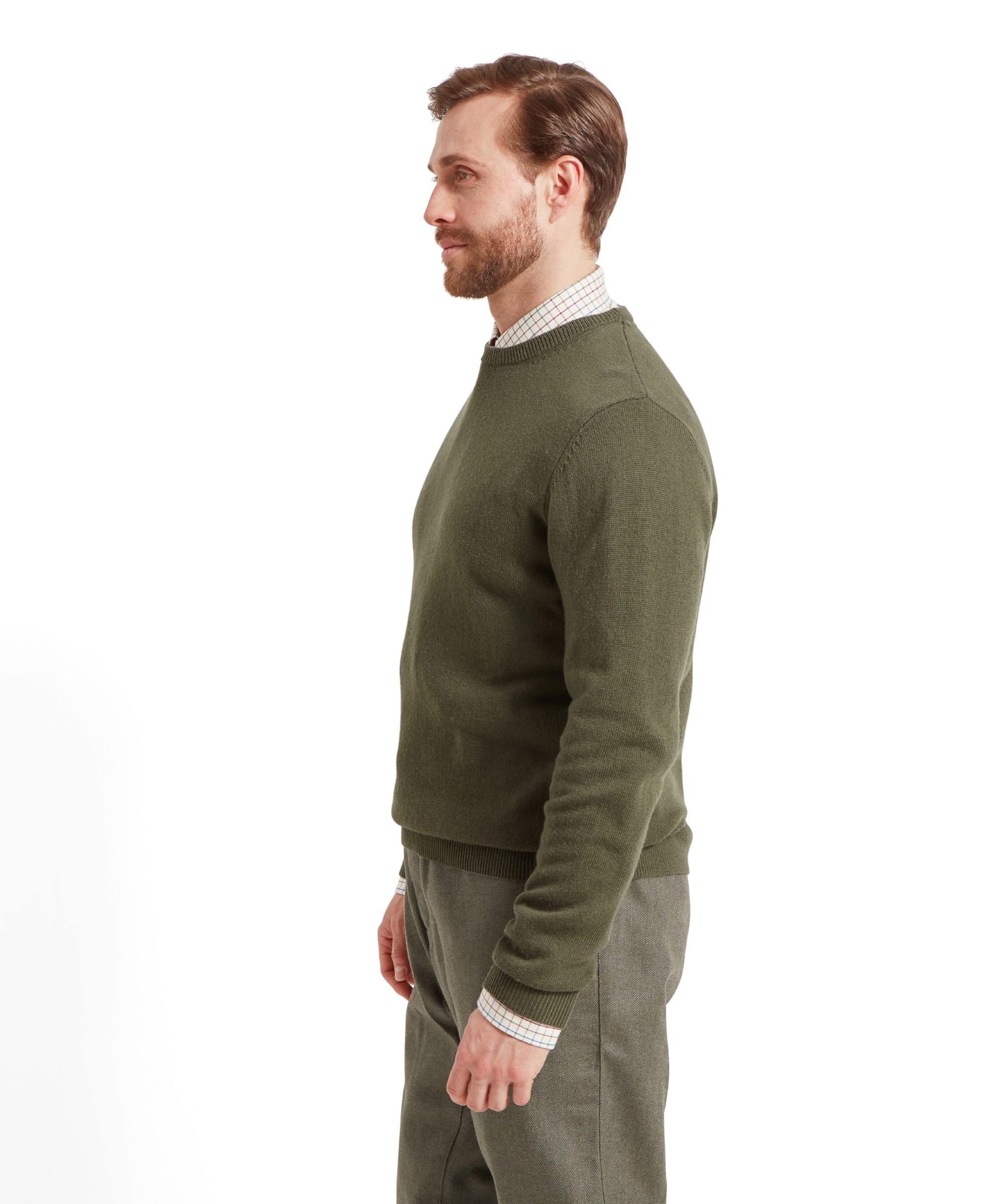 A man standing in profile against a plain white background, wearing a green crew‑neck jumper layered over a checked shirt with the cuffs showing, paired with green tweed trousers.