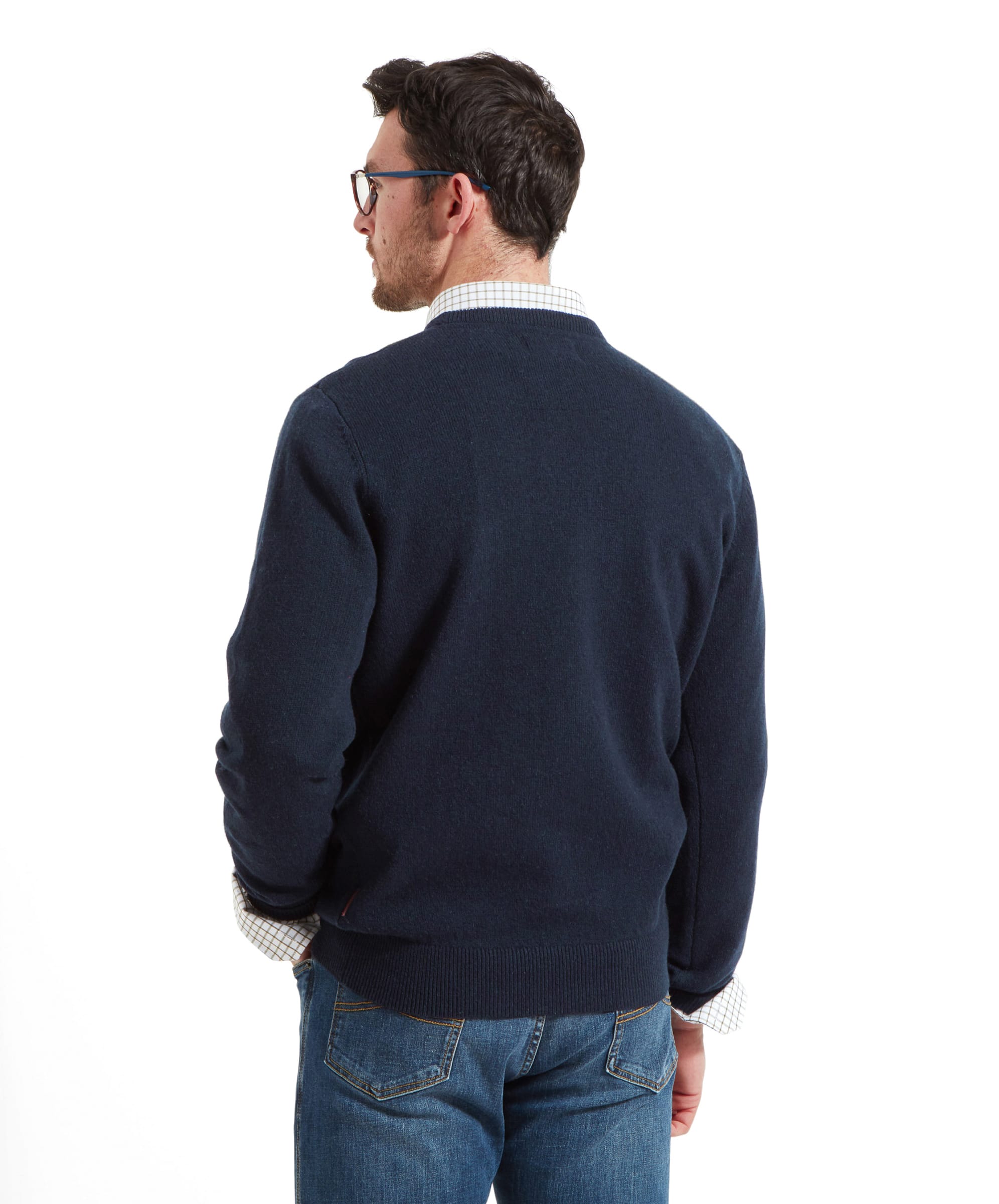 Back view of a man standing against a plain white background wearing a navy knitted jumper over a checked shirt, paired with blue jeans, with glasses visible and one hand resting by his side.