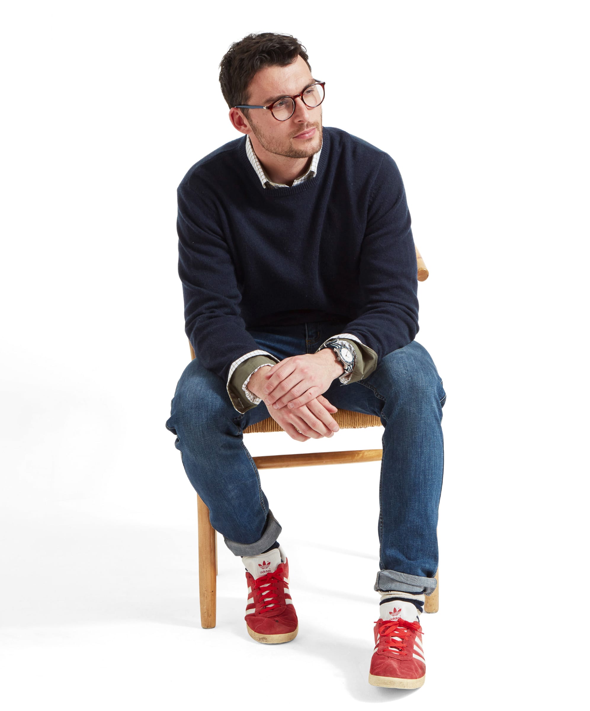 A man seated on a wooden chair against a plain white background wearing a navy knitted jumper over a checked shirt, blue jeans with rolled cuffs, patterned white socks and red trainers, with glasses and a wristwatch visible as he leans forward with hands clasped.