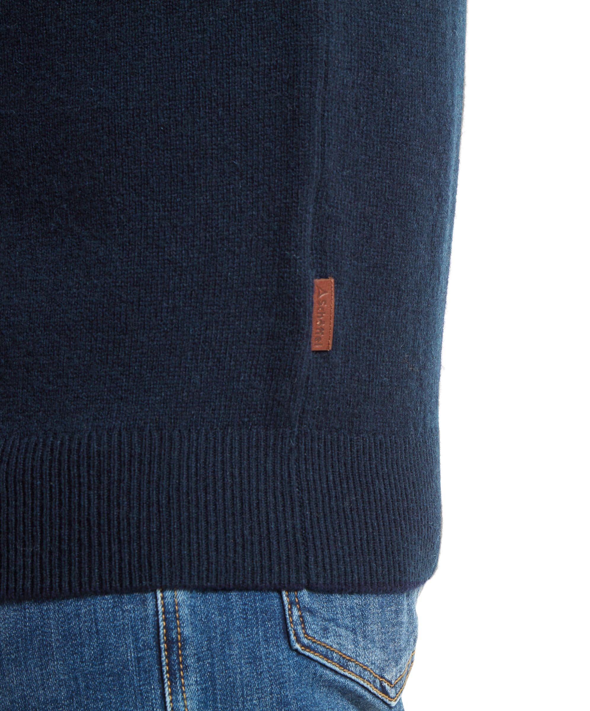 Close‑up of the hem of a navy knitted jumper showing a small brown branded tab, worn with blue denim jeans.