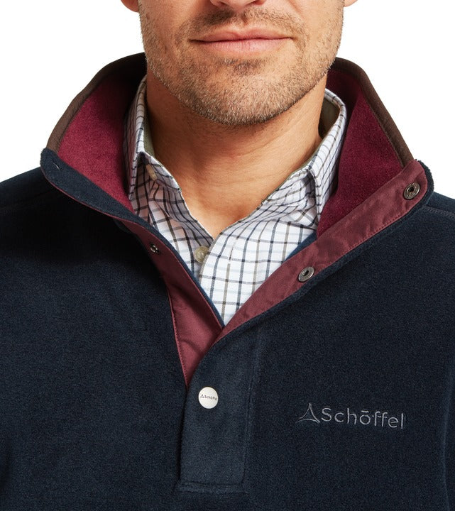 Schöffel Norton men's fleece in navy, close-up of high funnel neck with contrast burgundy trim, popper button fastening and embroidered logo, worn over checked shirt