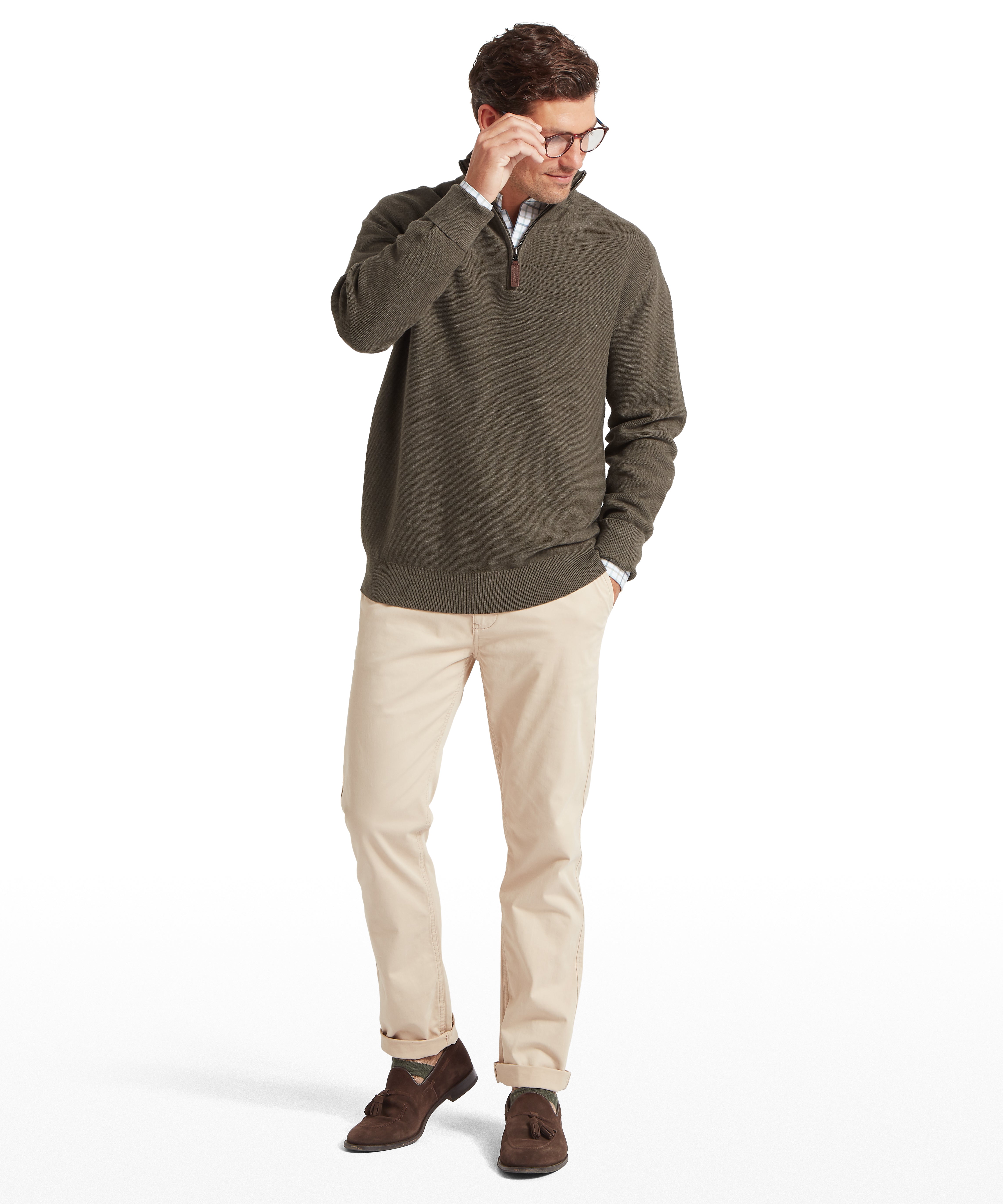 Man with glasses looking downwards, wearing a green jumper and beige trousers against a white background