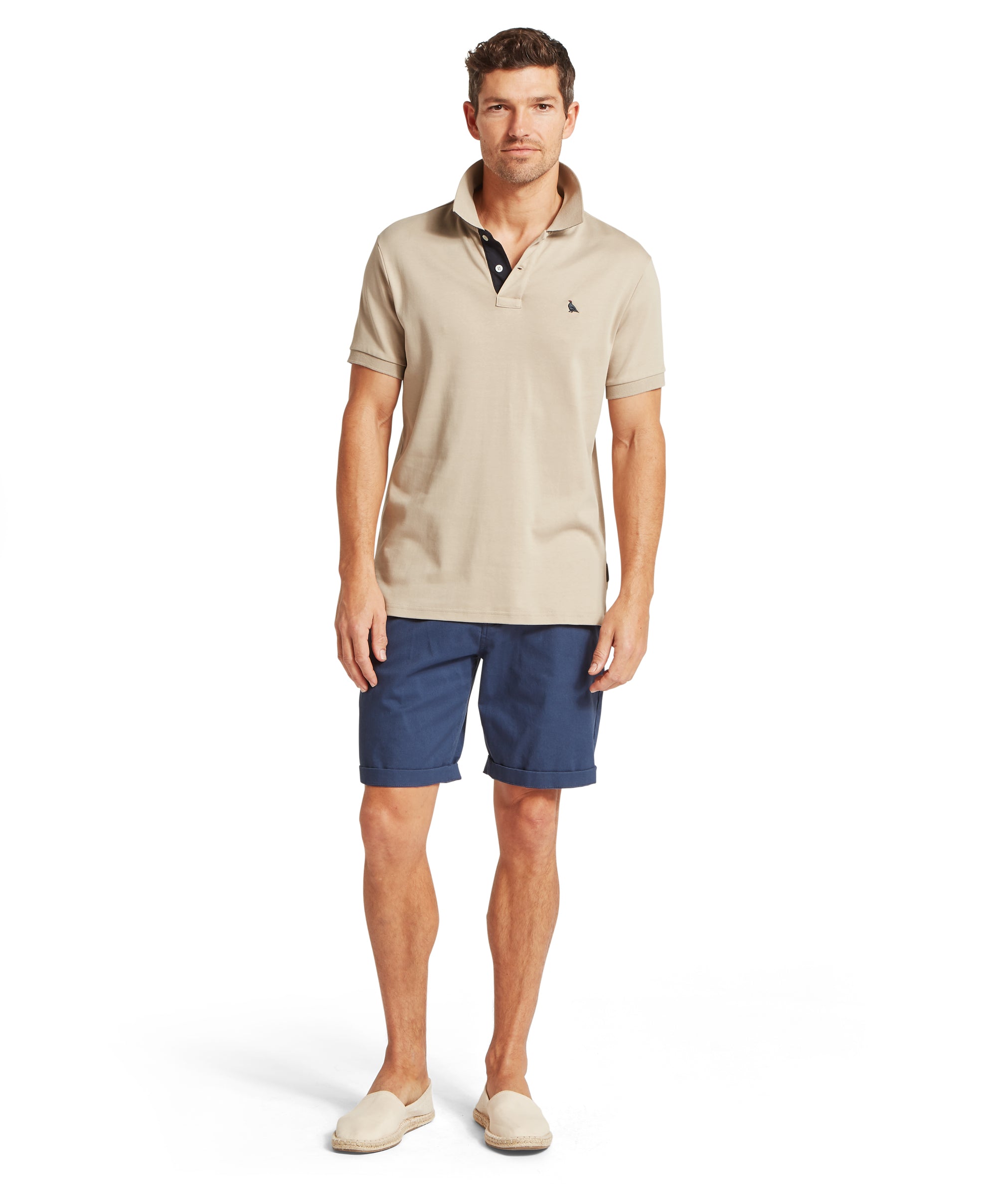 A man standing facing forward, wearing a beige short-sleeved polo shirt with a raised collar, blue tailored shorts and light slip-on espadrilles, photographed against a white background.