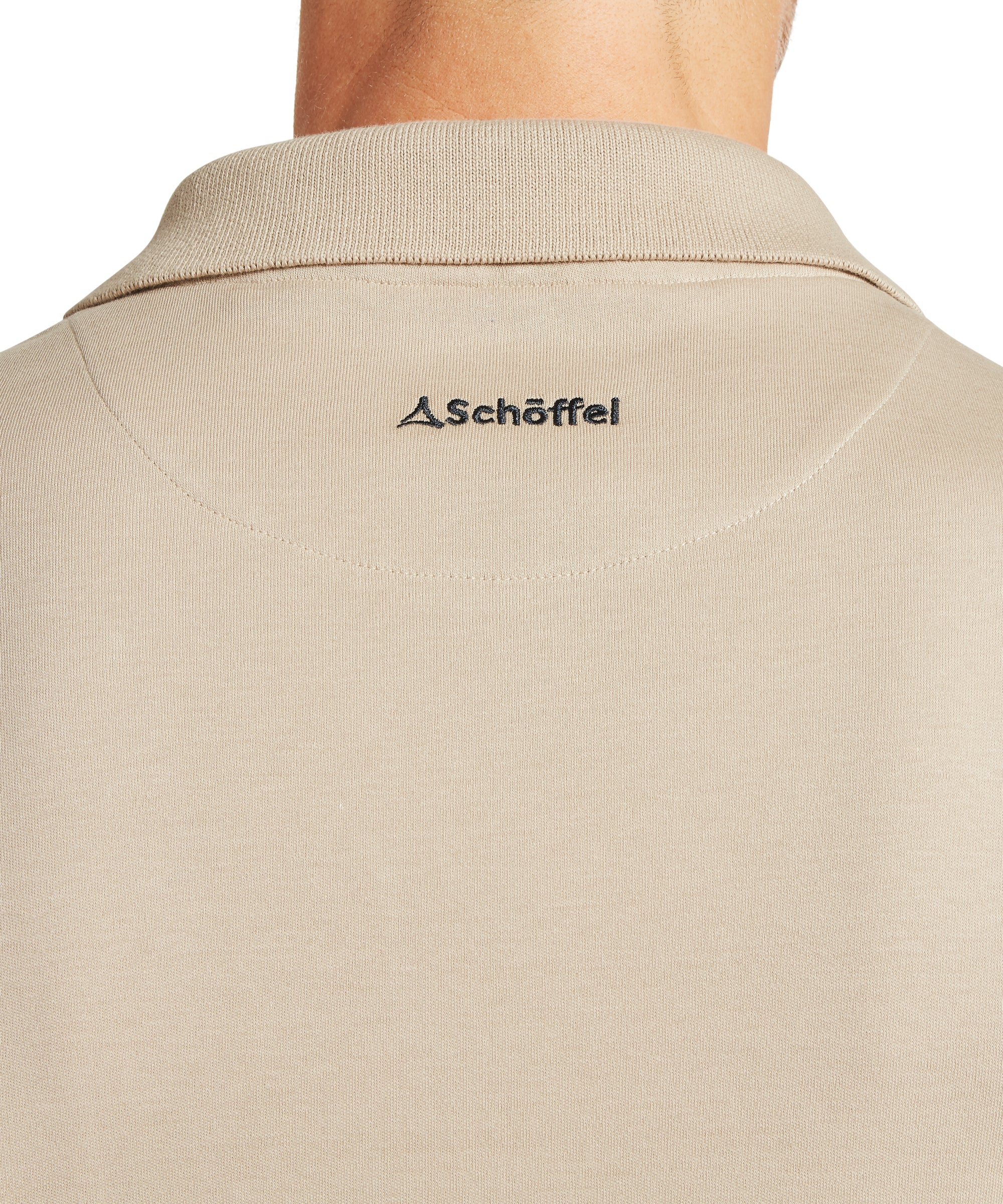 Close-up of the back of a man’s neck and upper back wearing a beige collared shirt. The shirt features the Schöffel logo embroidered below the collar, with a small triangular symbol to the left of the brand name. Branding detail shown in a neutral setting.