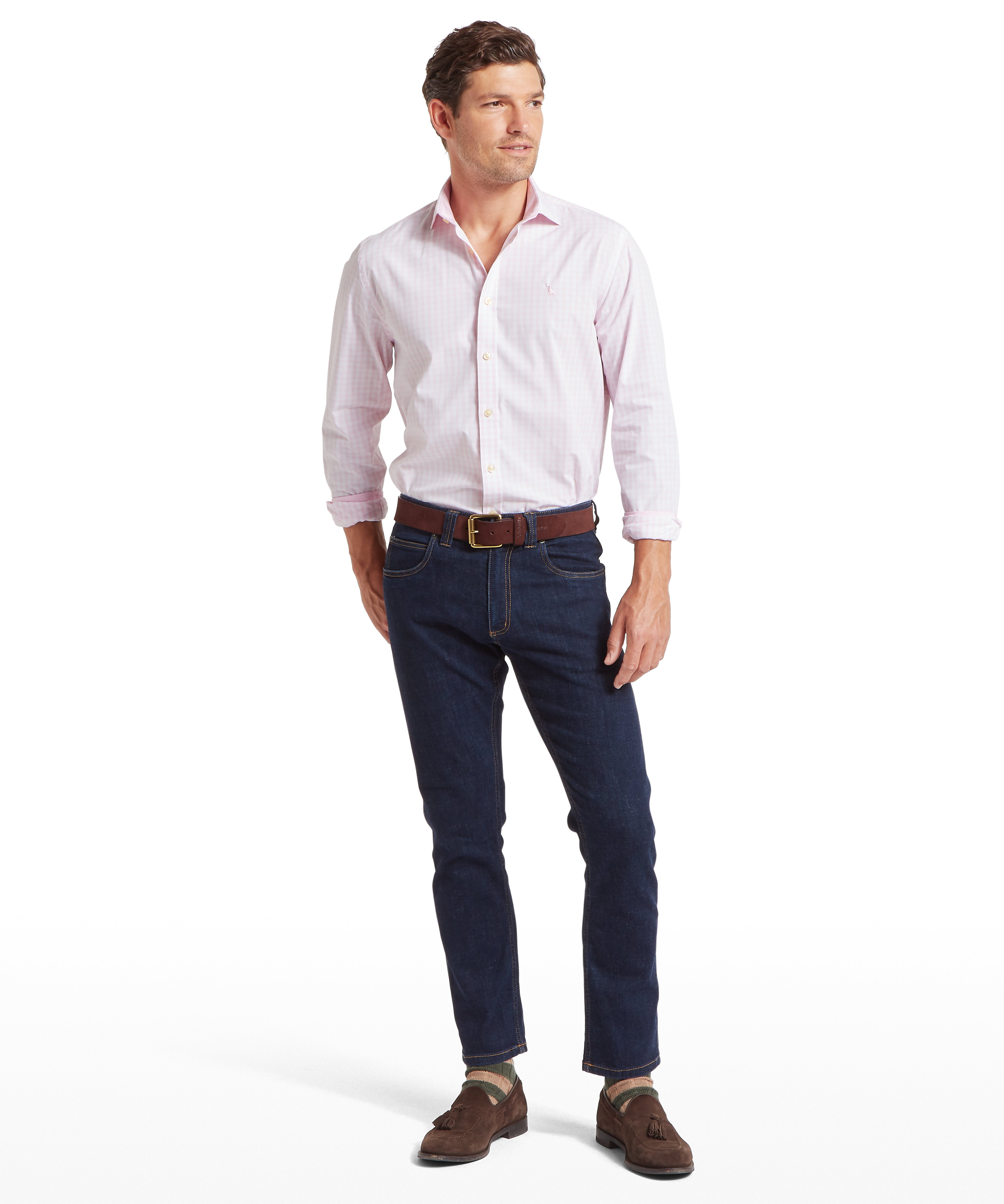 Man wearing a light pink shirt, dark blue jeans, and brown shoes against  a white background