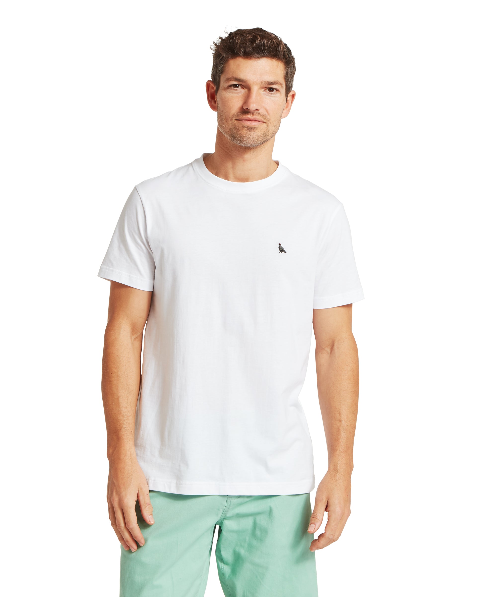 Man wearing a plain white short-sleeved T-shirt with a small black embroidered ptarmigan bird logo on the left chest. Paired with light green shorts. Standing front-facing in a relaxed pose against a plain white background.