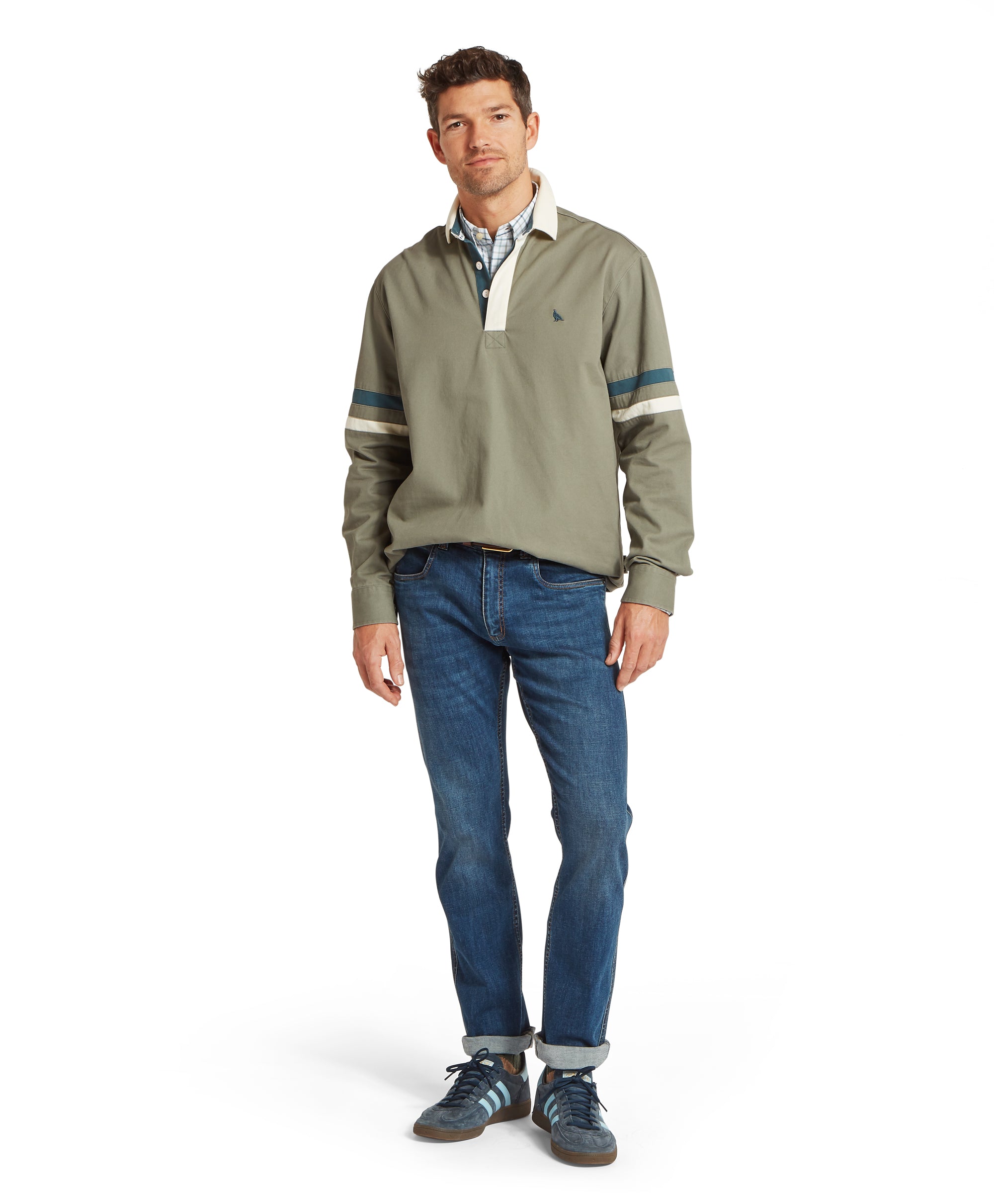 Man standing and facing forward wearing a light olive green deck shirt with a white collar and dark green and white sleeve stripes, layered over a blue and white checked shirt. Styled with medium-wash blue jeans with cuffed hems and dark trainers with blue stripes. The outfit presents a relaxed, everyday look with coordinated colour