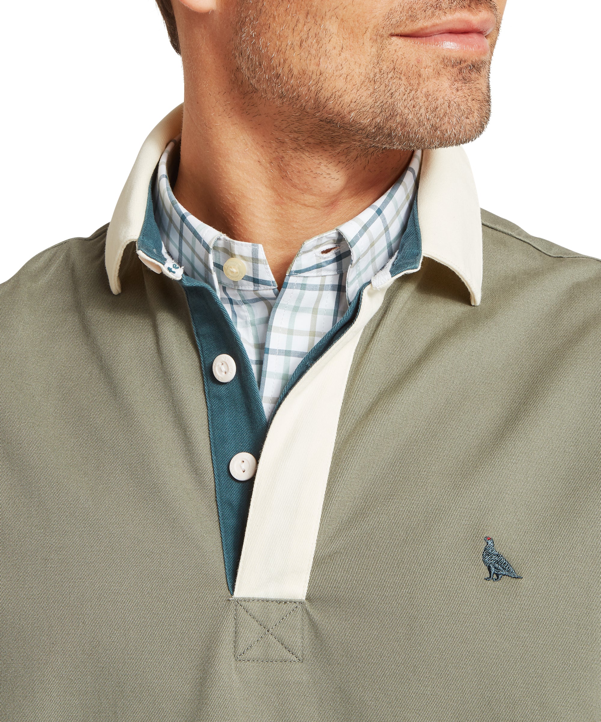 Close-up of a man’s upper torso wearing a green deck shirt with a white collar and teal placket, layered over a light blue and white checked button-down shirt. The deck shirt features a small embroidered ptarmigan bird logo on the left chest. The collar is turned up, revealing the shirt collar and buttons beneath.