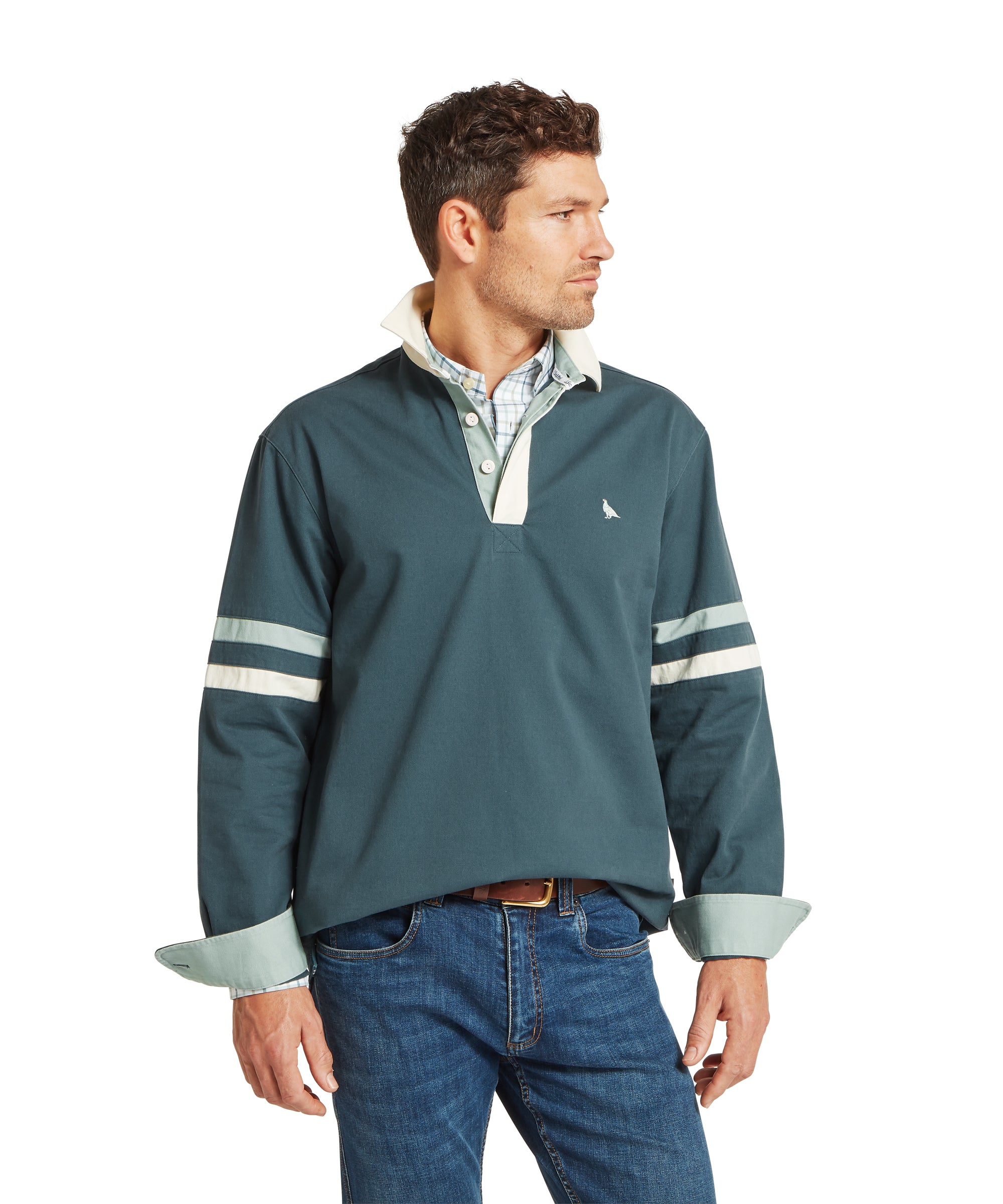 Man wearing a dark teal rugby-style jumper with white sleeve stripes and a small white chest logo, layered over a light checkered collared shirt. Styled with blue jeans and a brown belt. He is standing and looking to his left, showcasing a casual, layered outfit suitable for cooler weather.