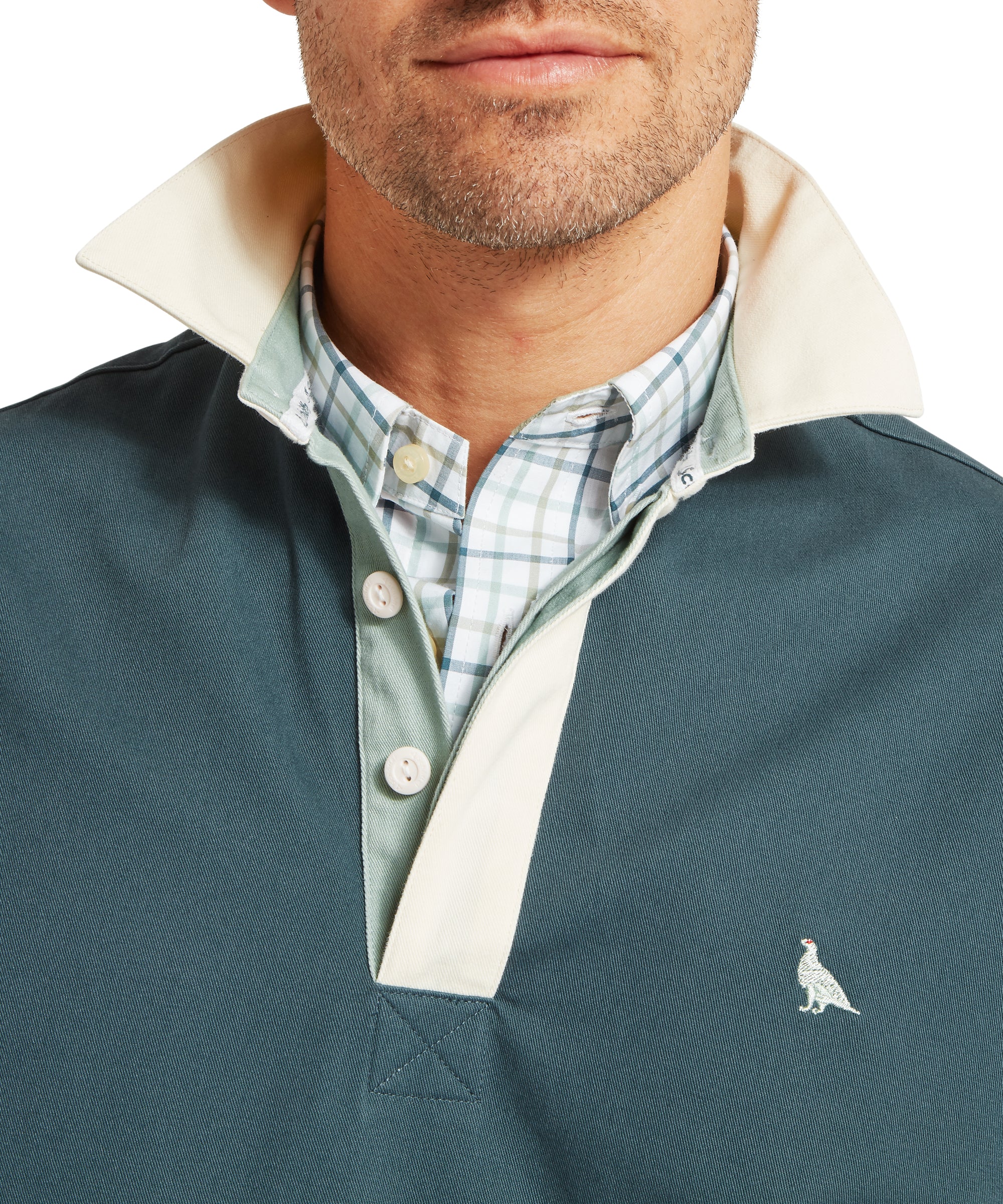 Close-up of a man’s upper torso wearing a teal deck shirt with a white collar and light green placket, layered over a blue and green checked button-down shirt. The deck shirt features a small embroidered bird logo on the left chest. The collar is turned up, revealing the shirt collar and buttons beneath.