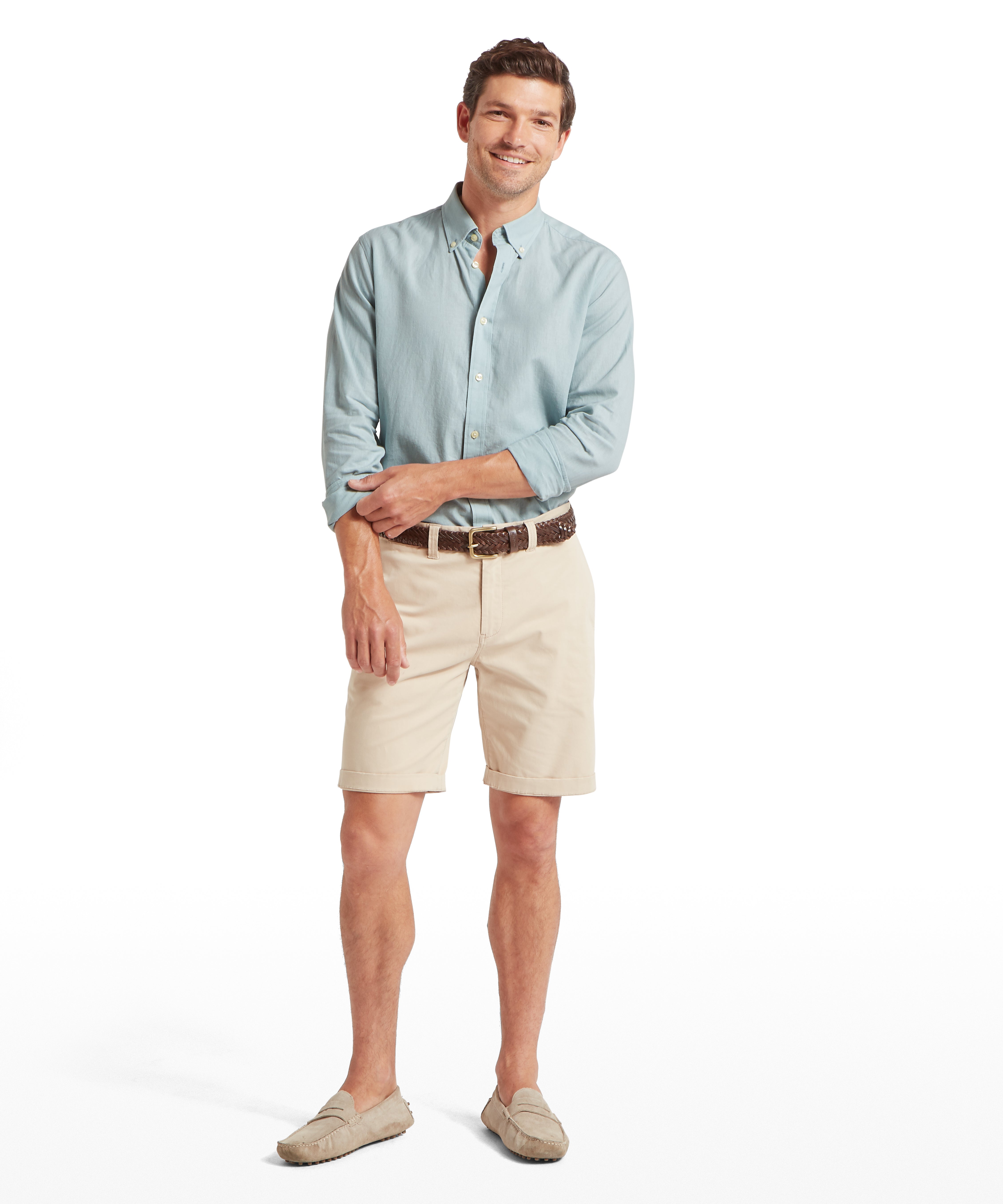 A man is standing against a plain white background, facing forward. He is wearing a long-sleeved blue linen shirt with a button-down collar, and the sleeves are rolled up to the mid-forearm. He is also wearing light beige chino shorts secured with a dark brown woven leather belt and beige suede loafers. He is smiling warmly at the camera.