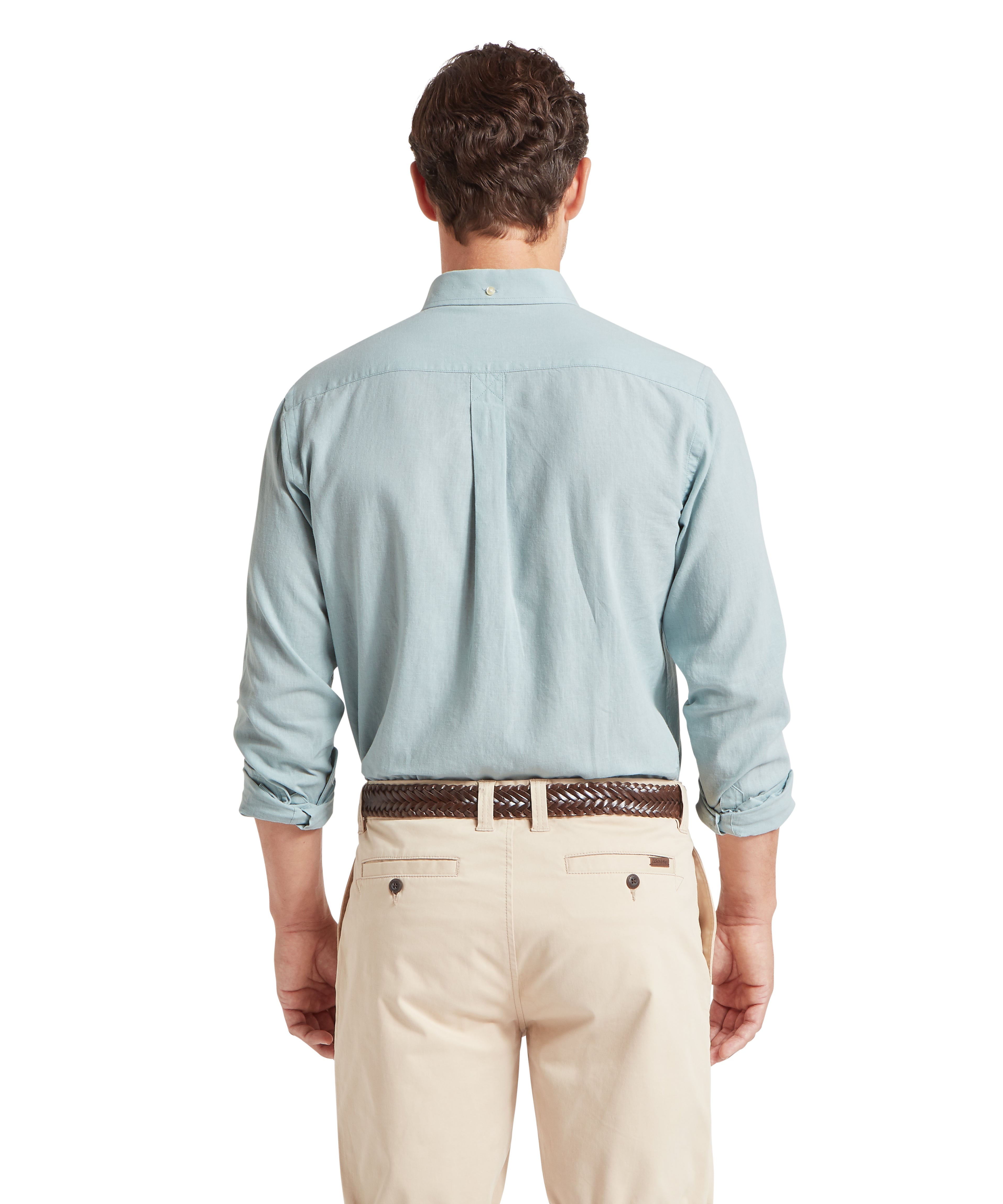 A rear view of a man with short, dark wavy hair. He is wearing a light blue long-sleeved button-down shirt with the sleeves rolled up to the elbows. The shirt is tucked into beige chinos, secured with a dark brown braided leather belt. The background is solid white.