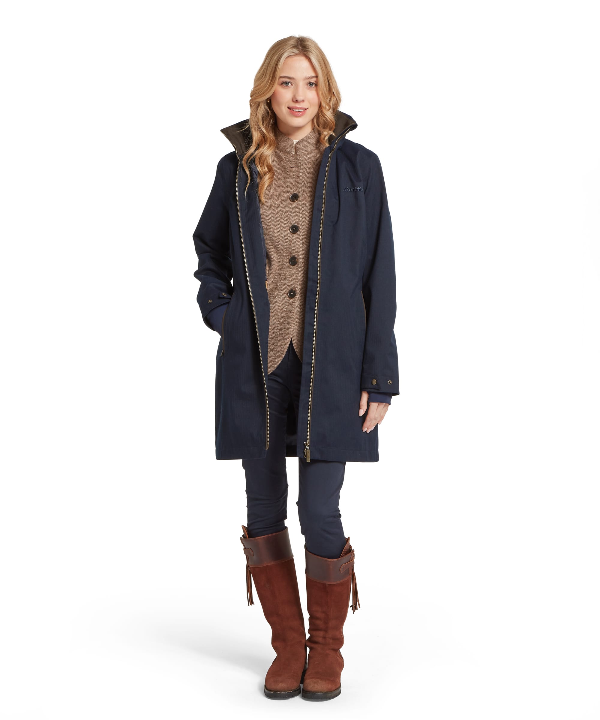 Full-length view of the Women's Schöffel Allexton Waterproof Coat in midnight navy, worn open over a camel tweed gilet and slim navy trousers, with tall tan suede and leather riding boots, showing the mid-length silhouette and antique brass zip.