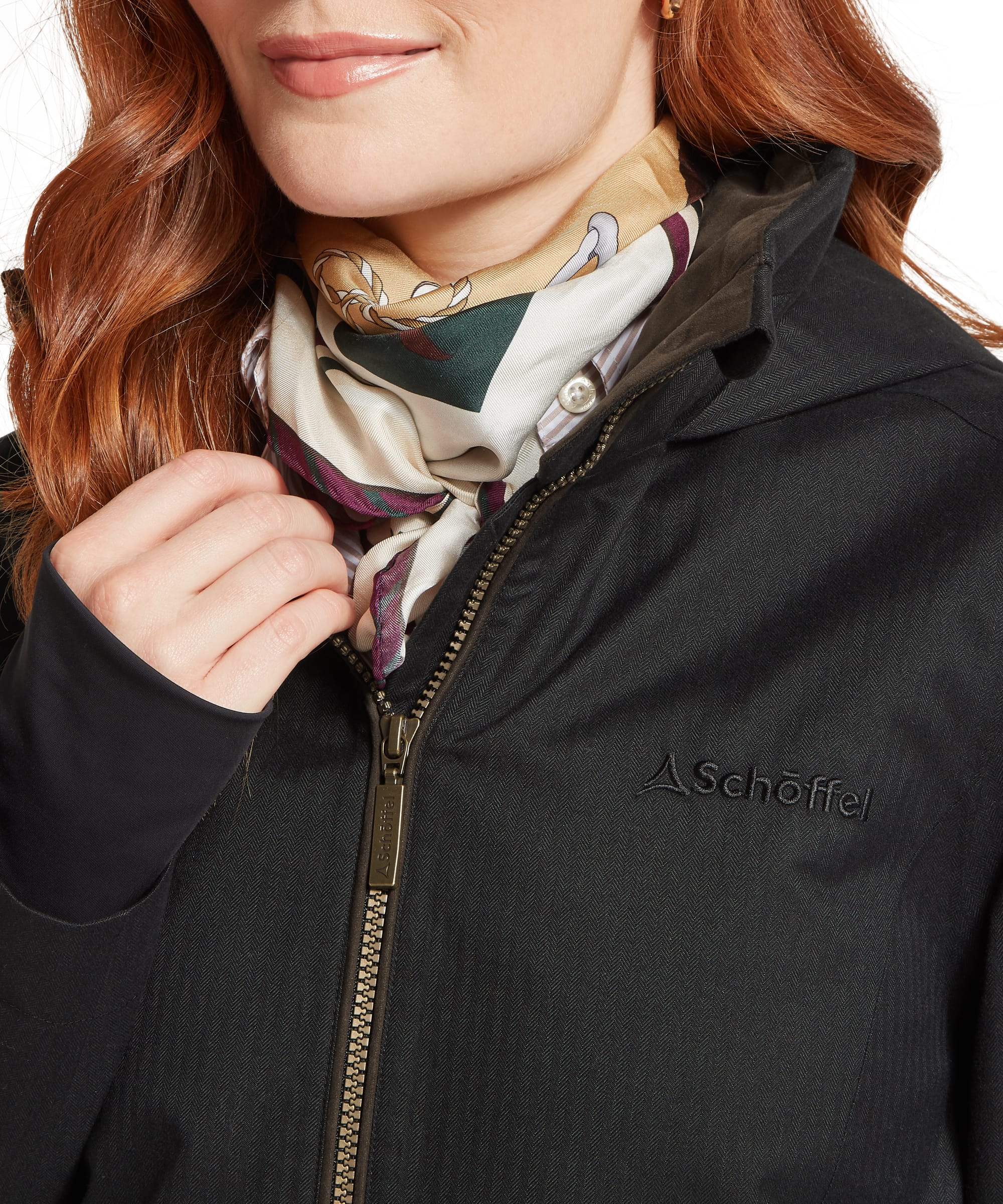 Close-up detail of the Women's Schöffel Allexton Waterproof Coat in charcoal herringbone, highlighting the tonal embroidered Schöffel logo, antique brass zip and inner storm collar, styled with a colourful silk scarf.