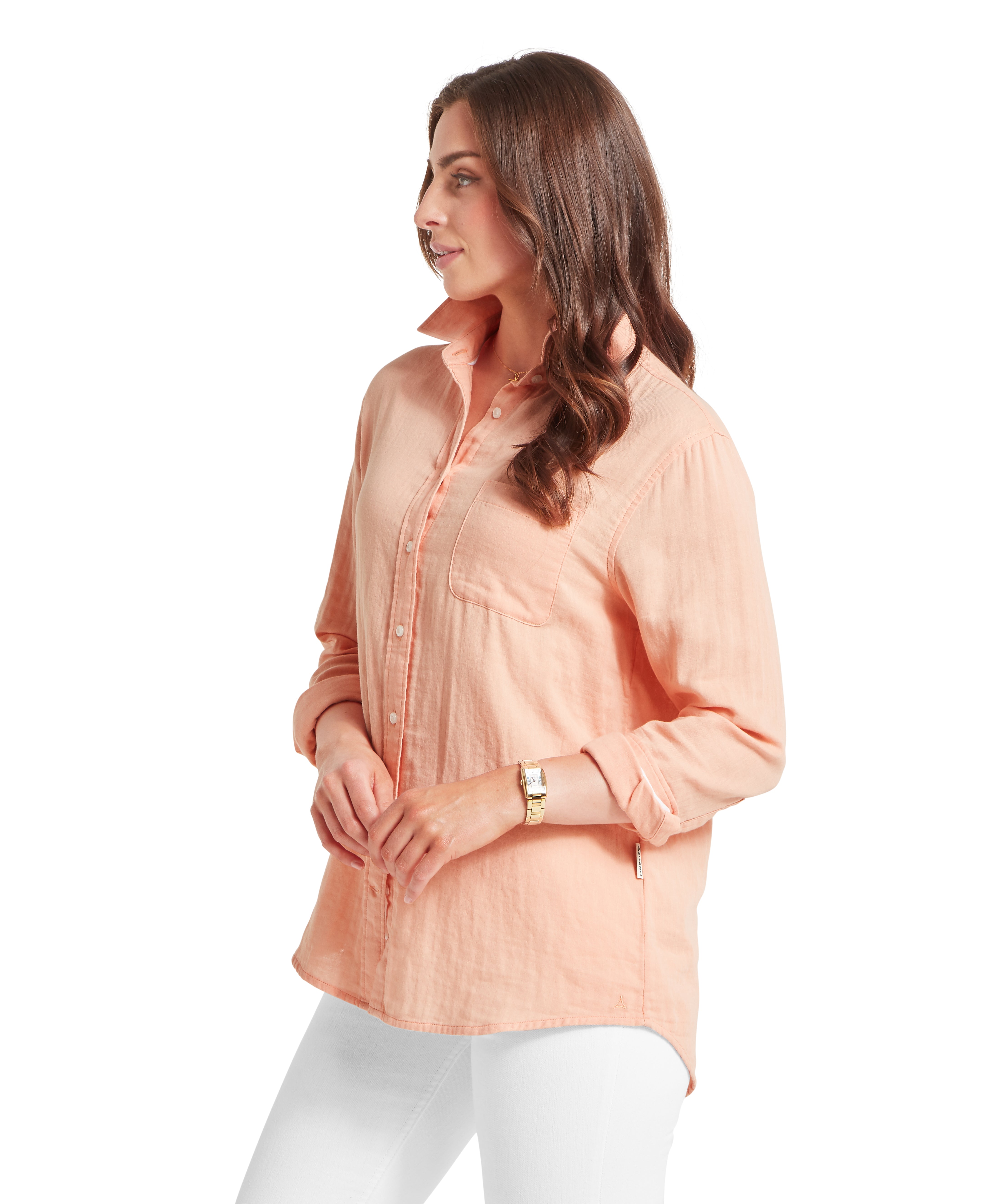 Woman standing in a side profile wearing a peach-coloured long-sleeved linen shirt with the sleeves loosely rolled up to the forearms, revealing a gold watch on her left wrist. The shirt is layered over white skinny jeans and features a visible chest pocket and a curved hemline. Her long brown hair is styled in soft waves and draped over her shoulder, complementing the relaxed look of the outfit.