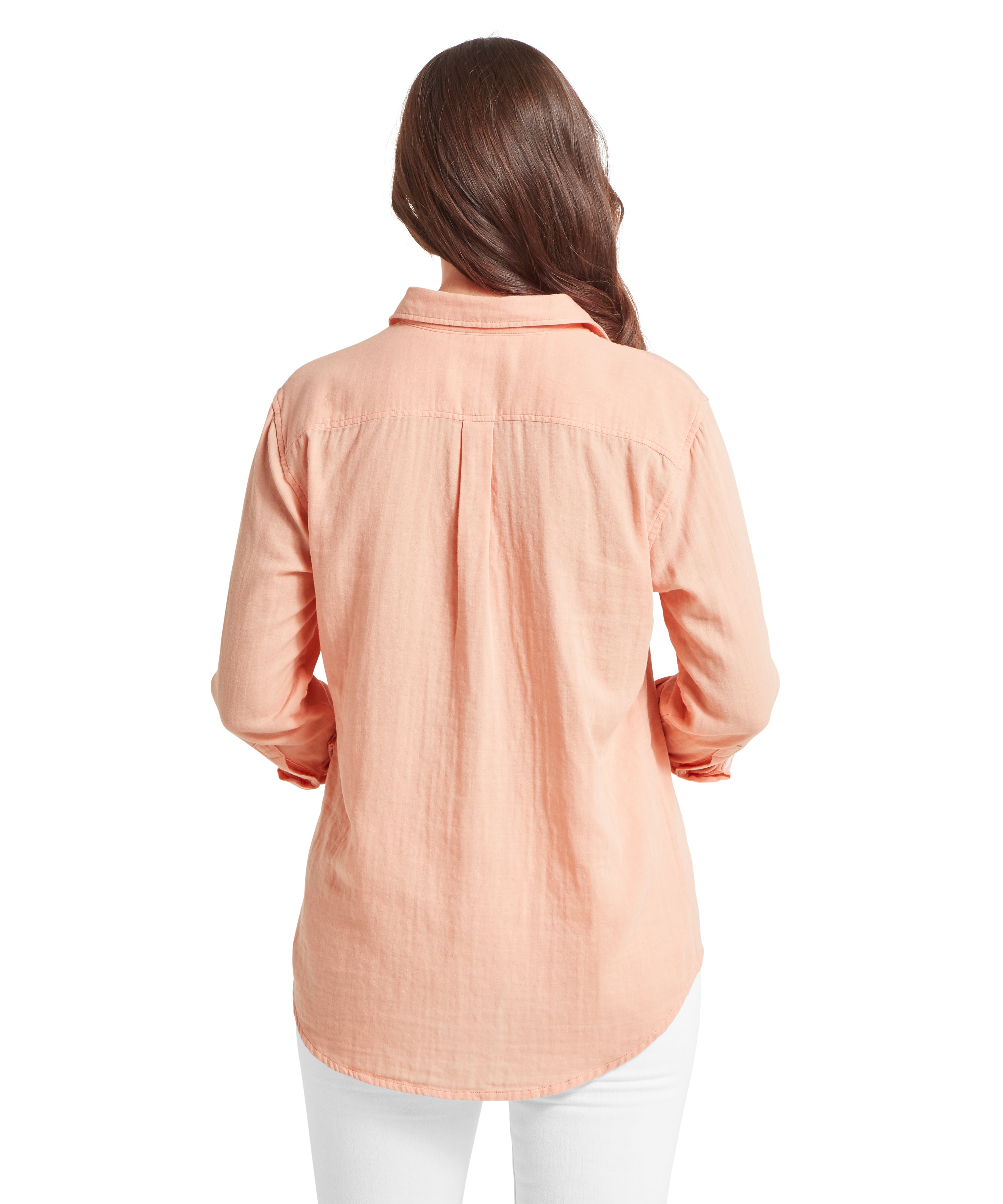 Woman seen from behind wearing a peach-coloured long-sleeved shirt, which features a back yoke with a central box pleat for a comfortable, relaxed fit. The shirt has a curved hemline and is worn over white trousers, with her long brown hair falling naturally down her back.