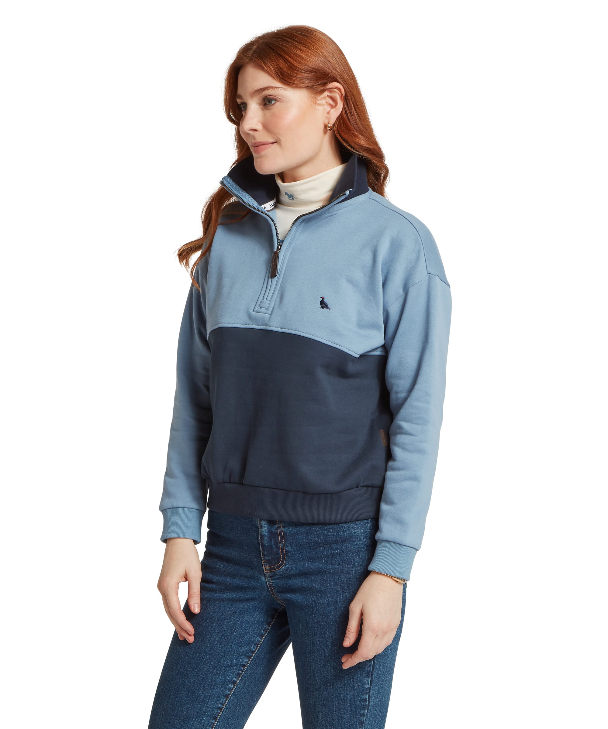 Three-quarter view of a woman wearing the Schöffel Women's Braunton 1/4 Zip Sweatshirt in two-tone Pale Blue and Navy, showing the colour-block design, embroidered Schöffel guinea fowl logo and quarter-zip placket, layered over a cream roll neck