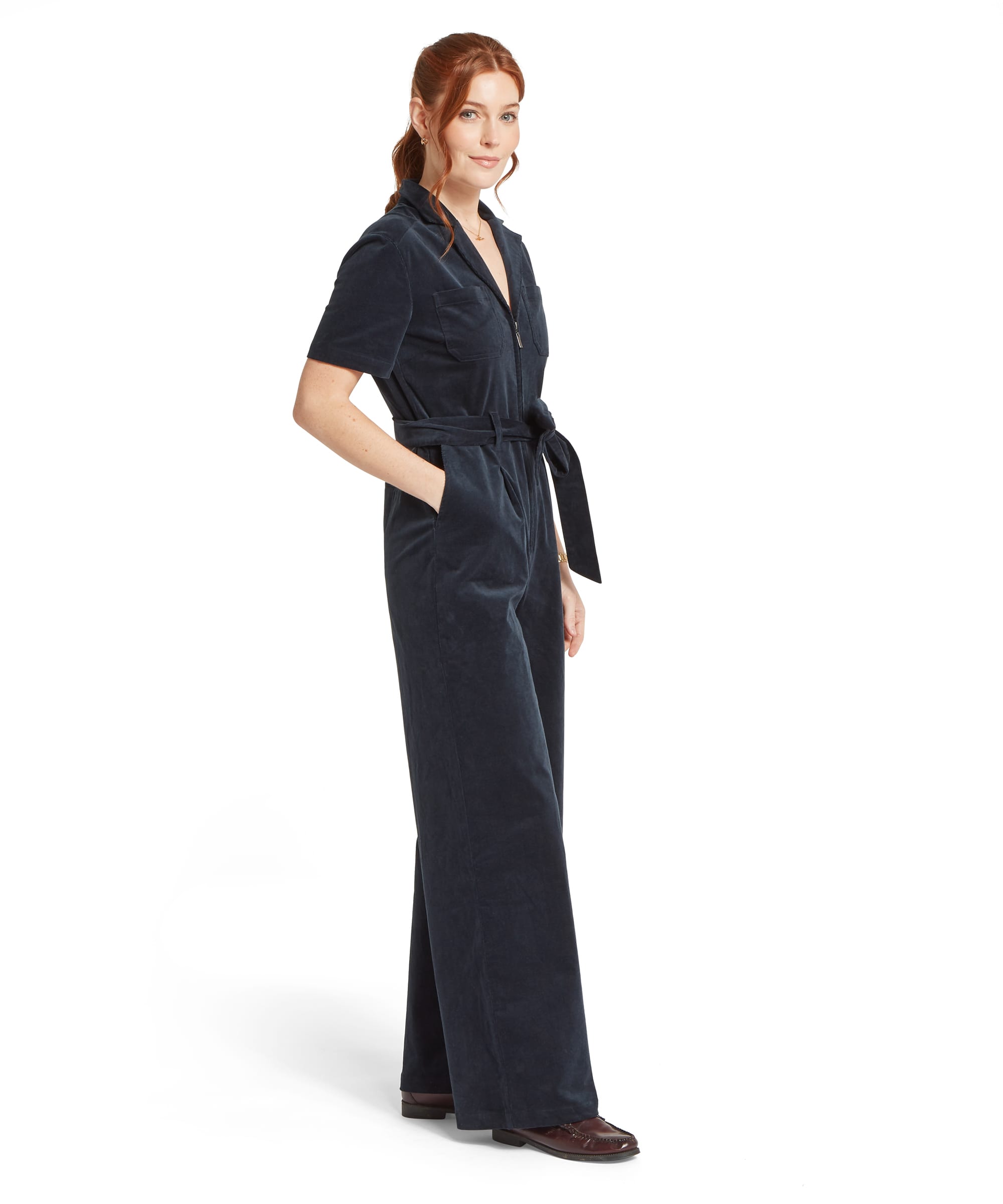 Side view of the Women's Schöffel Briar Cord Jumpsuit in midnight navy needlecord, featuring a V-neck with notched collar, short sleeves, self-tie waist belt and wide-leg trousers, worn with dark burgundy leather brogues.