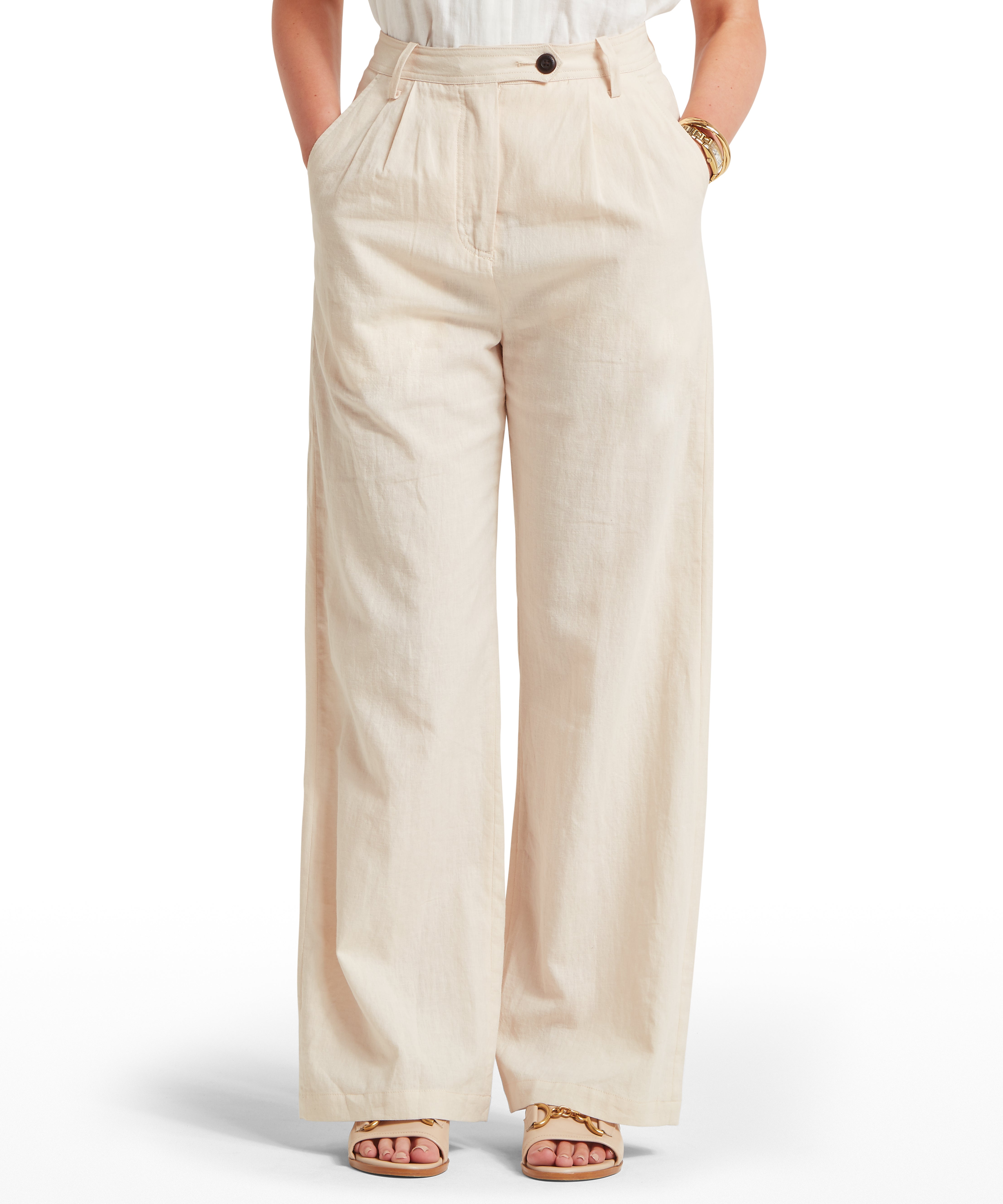Woman wearing high-waisted, wide-leg beige trousers with pleated front, side pockets, belt loops, and button fastening. Paired with a sleeveless tucked-in top and open-toe sandals. Plain white background highlights the outfit’s lightweight fabric and relaxed styling.