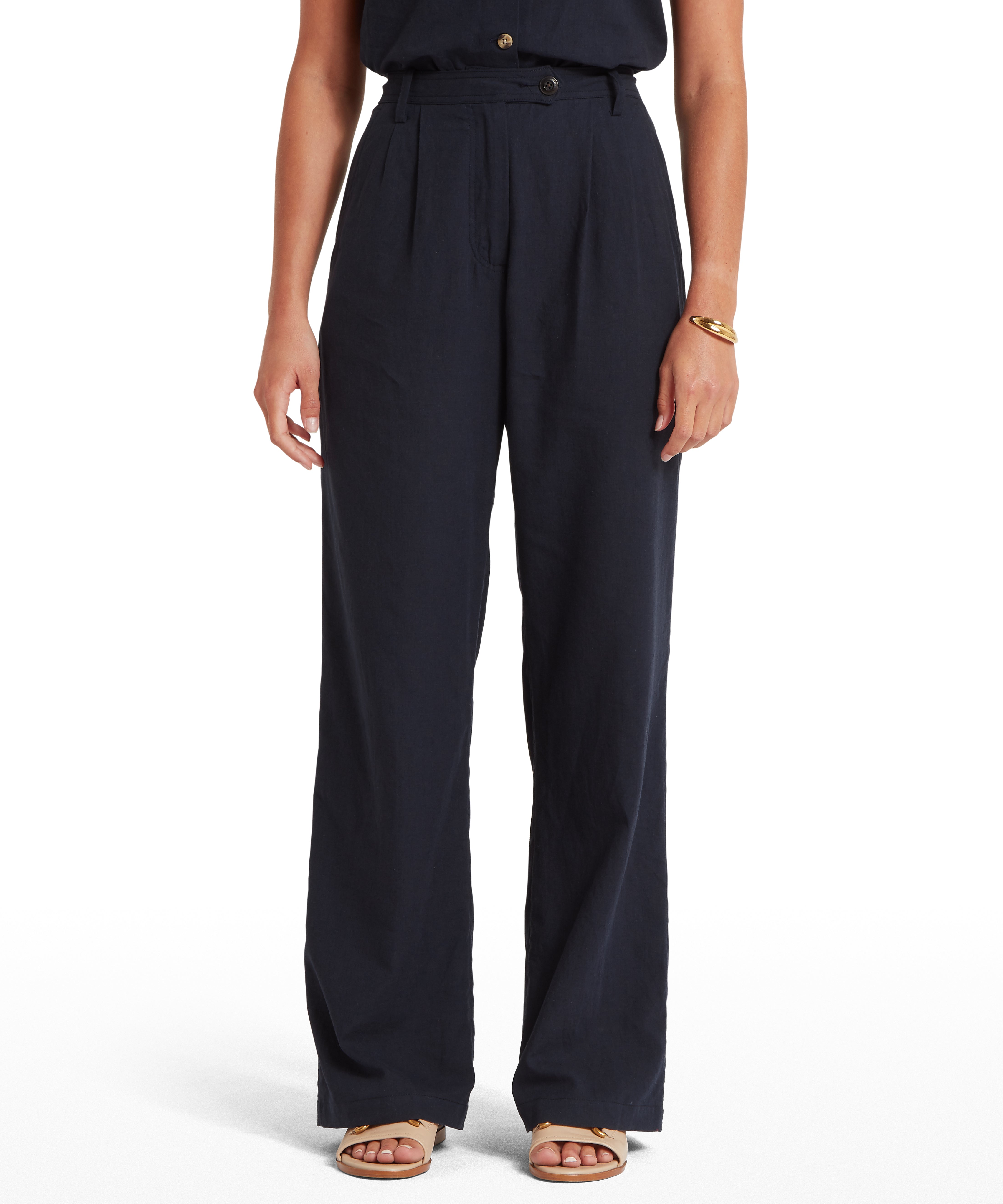 Woman wearing high-waisted, wide-leg navy trousers with front pleats, belt and button fastening. Paired with a sleeveless tucked-in top, gold bracelet on right wrist, and open-toe sandals. Plain white background keeps focus on the outfit’s silhouette and styling.