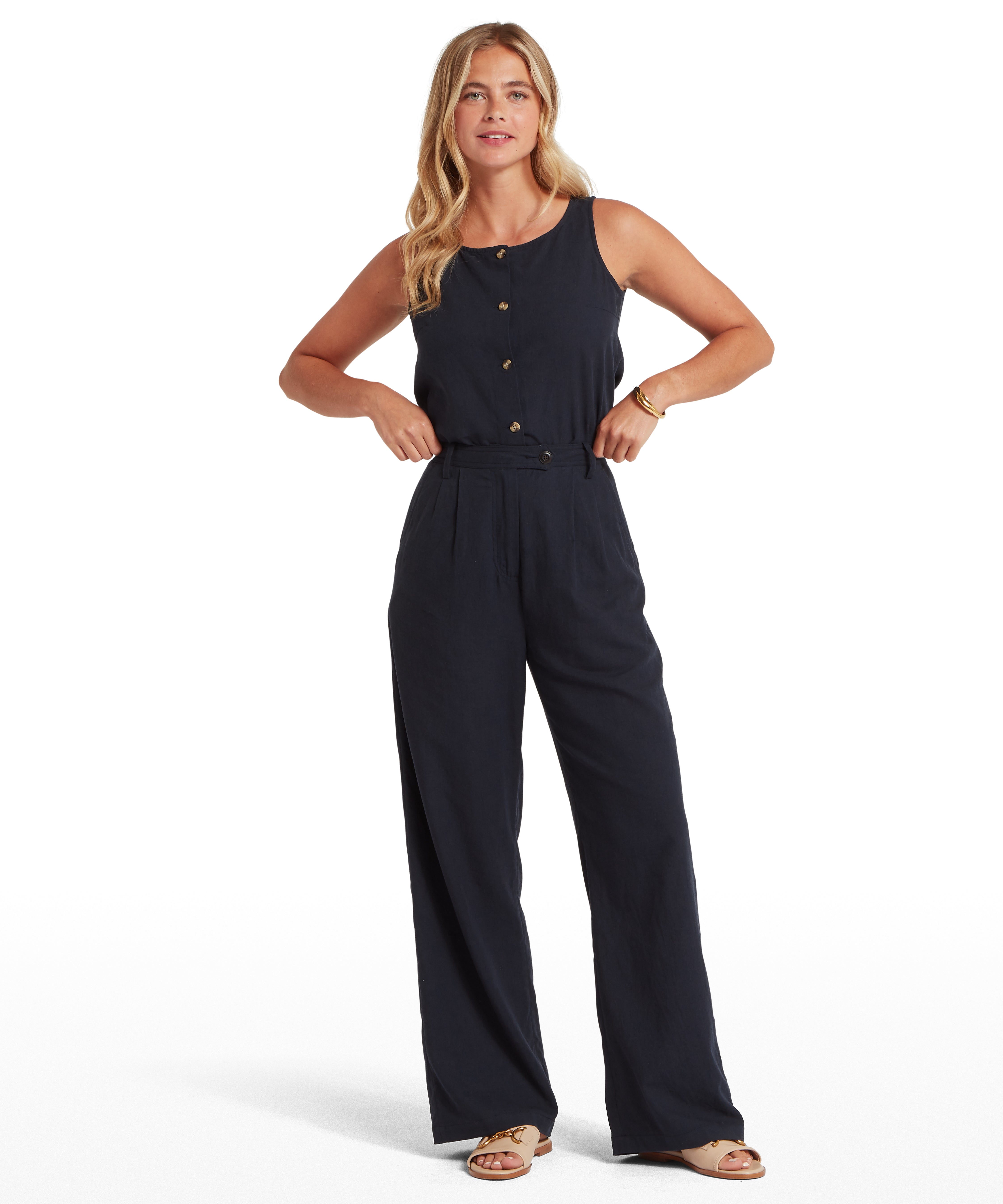 Woman standing in a sleeveless navy co-ord featuring a sleeveless vest top and wide-leg trousers.  Styled with flat sandals and a wristwatch. Long, wavy blonde hair visible. Plain white background keeps focus on the outfit’s fit and detailing.