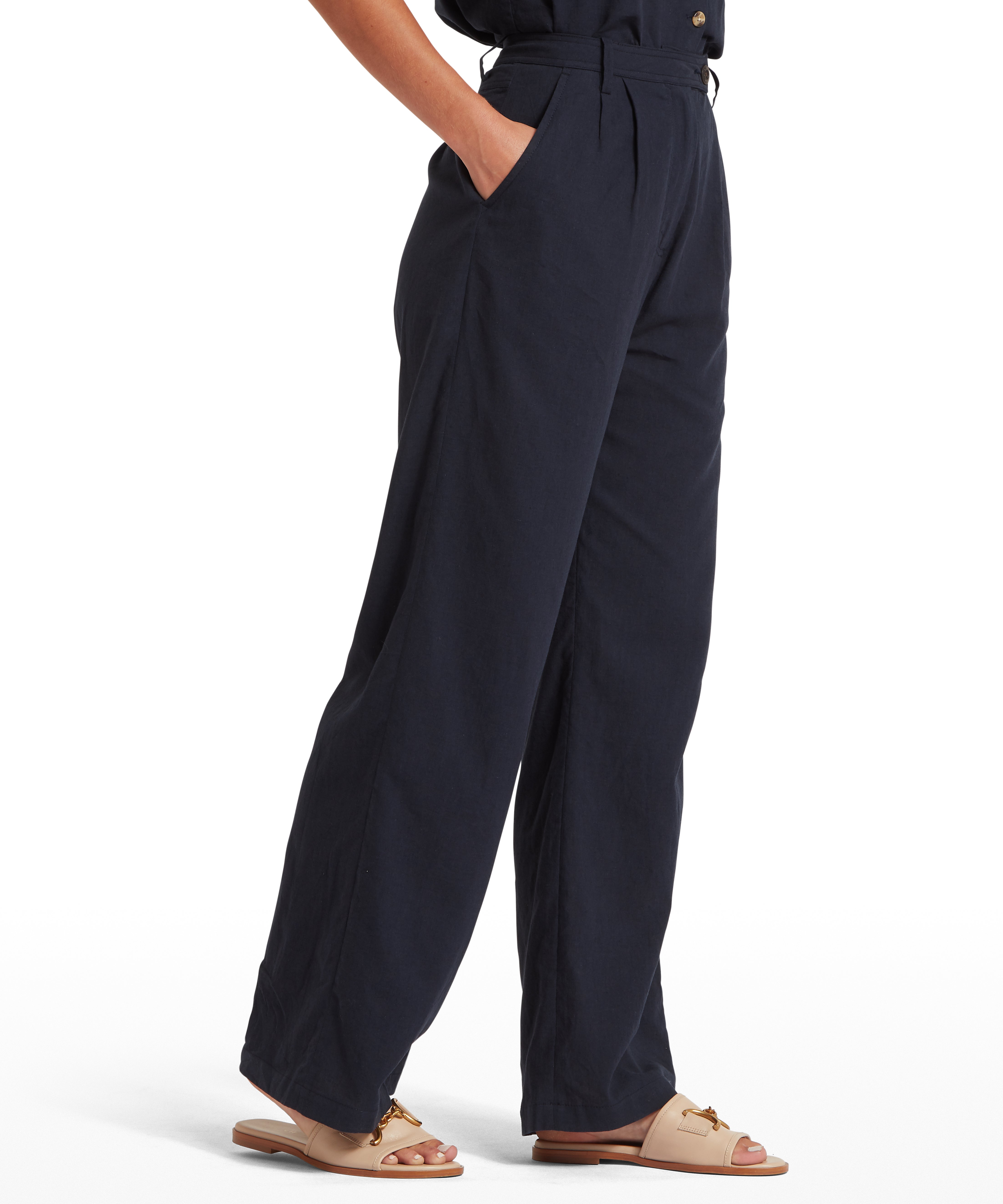 Woman wearing high-waisted, wide-leg navy trousers with pleated waist and side pockets. Paired with beige flat sandals featuring gold chain detail. Standing with one hand in pocket. Plain white background highlights the fit and styling of the trousers.