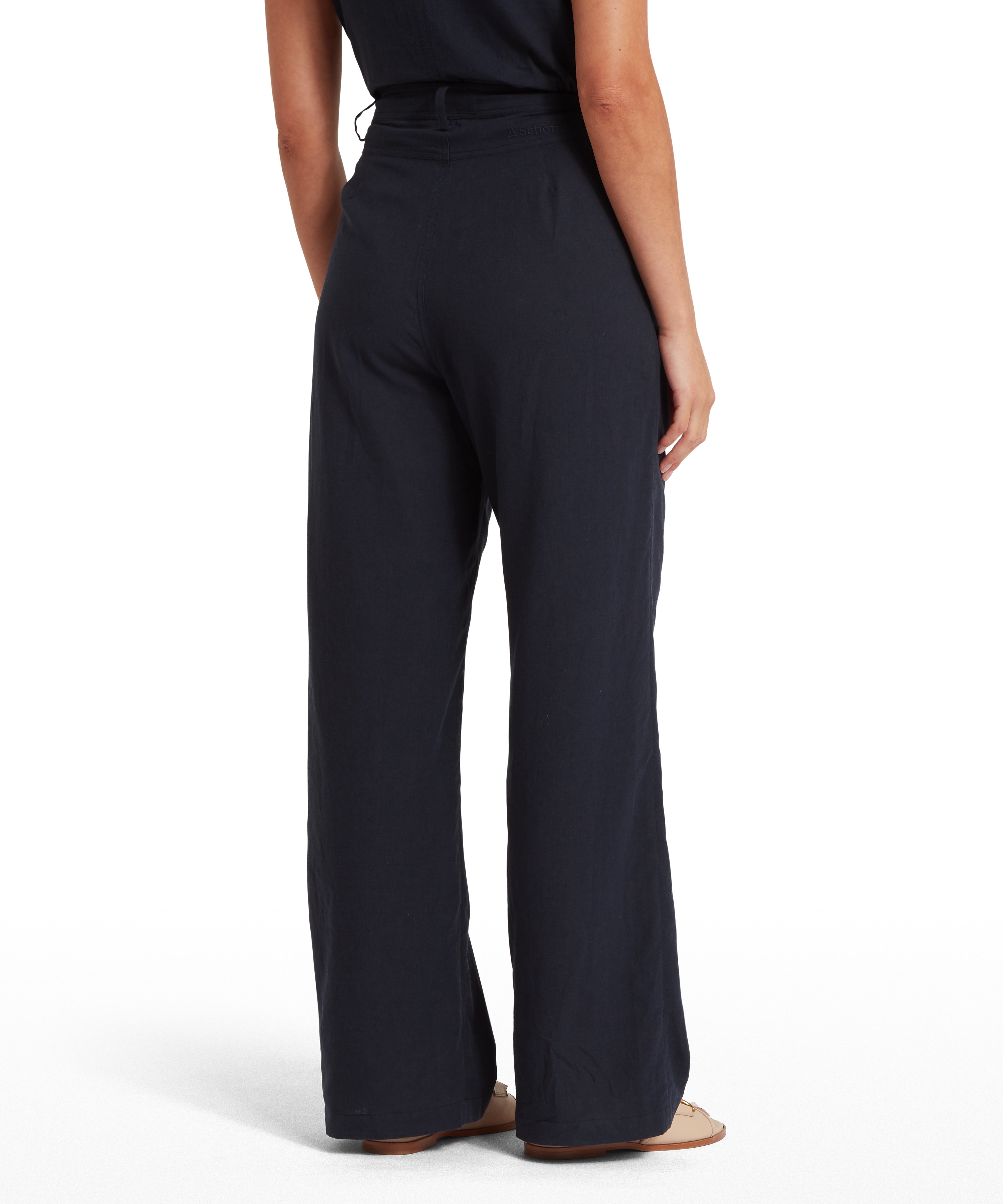 Rear view of a woman wearing dark wide-leg trousers and a matching sleeveless top. Trousers have a high waist and loose fit, paired with light-coloured flat sandals. Plain white background draws attention to the silhouette and garment fit.