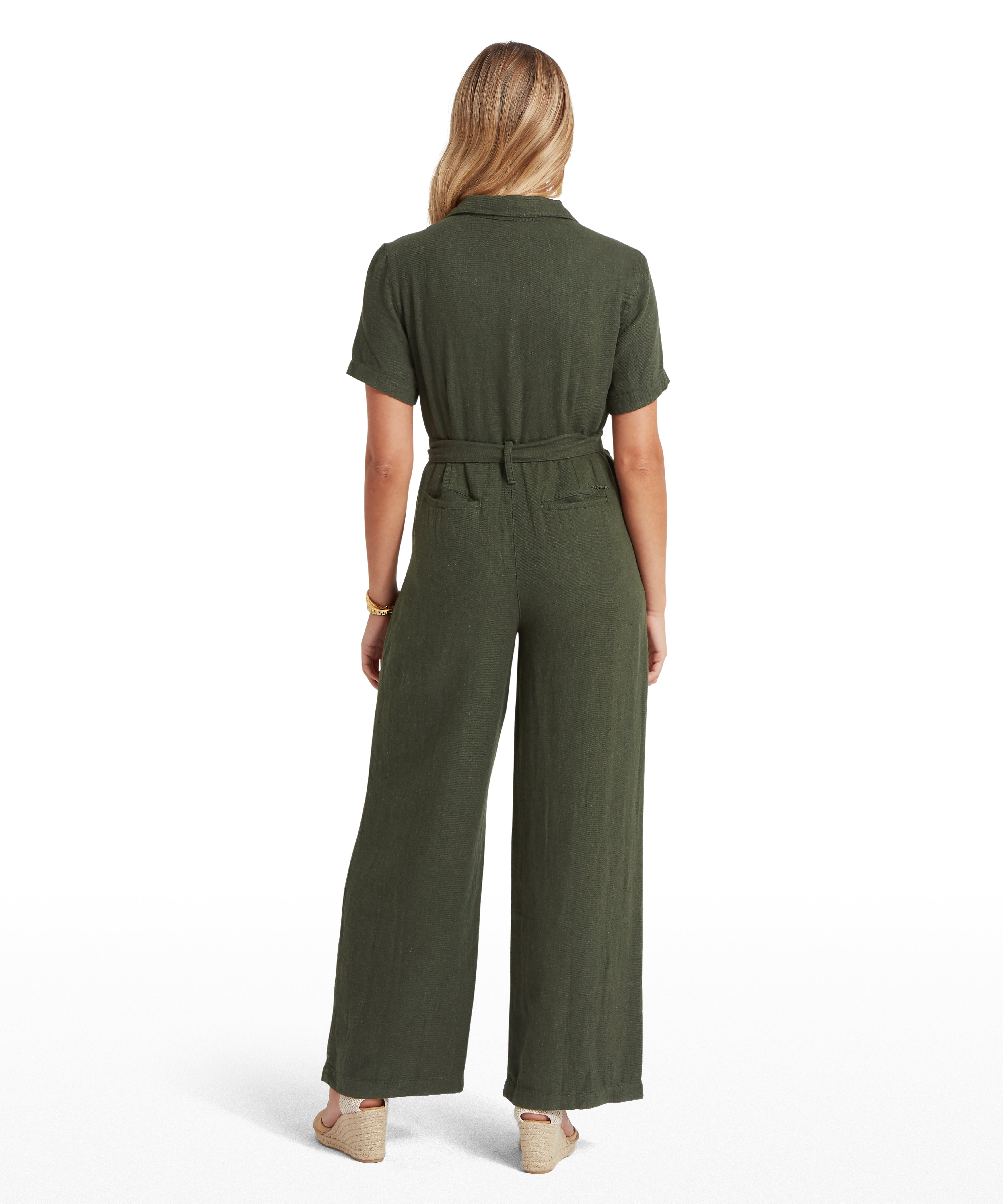 Back view of a woman wearing a dark green short-sleeved jumpsuit with a belt tied at the waist. The jumpsuit has a textured fabric and wide-leg trousers that reach the ankles. She is also wearing wedge sandals and a bracelet on her right wrist. Displayed against a plain white background.