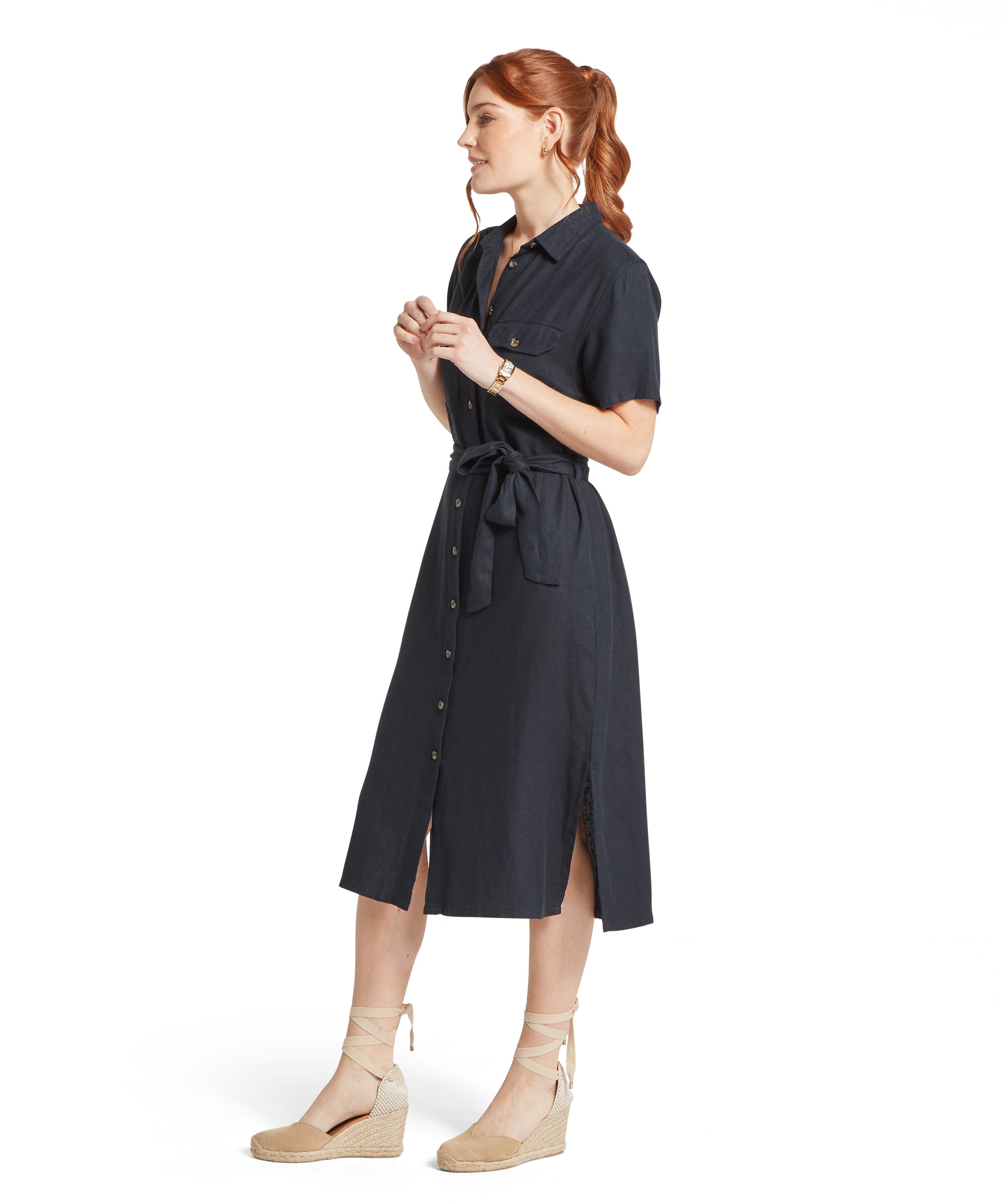 A woman standing against a plain white background wearing a navy linen button‑down midi dress with short sleeves, a waist tie, a front slit and a chest pocket, paired with beige espadrille wedge sandals with ankle ties.