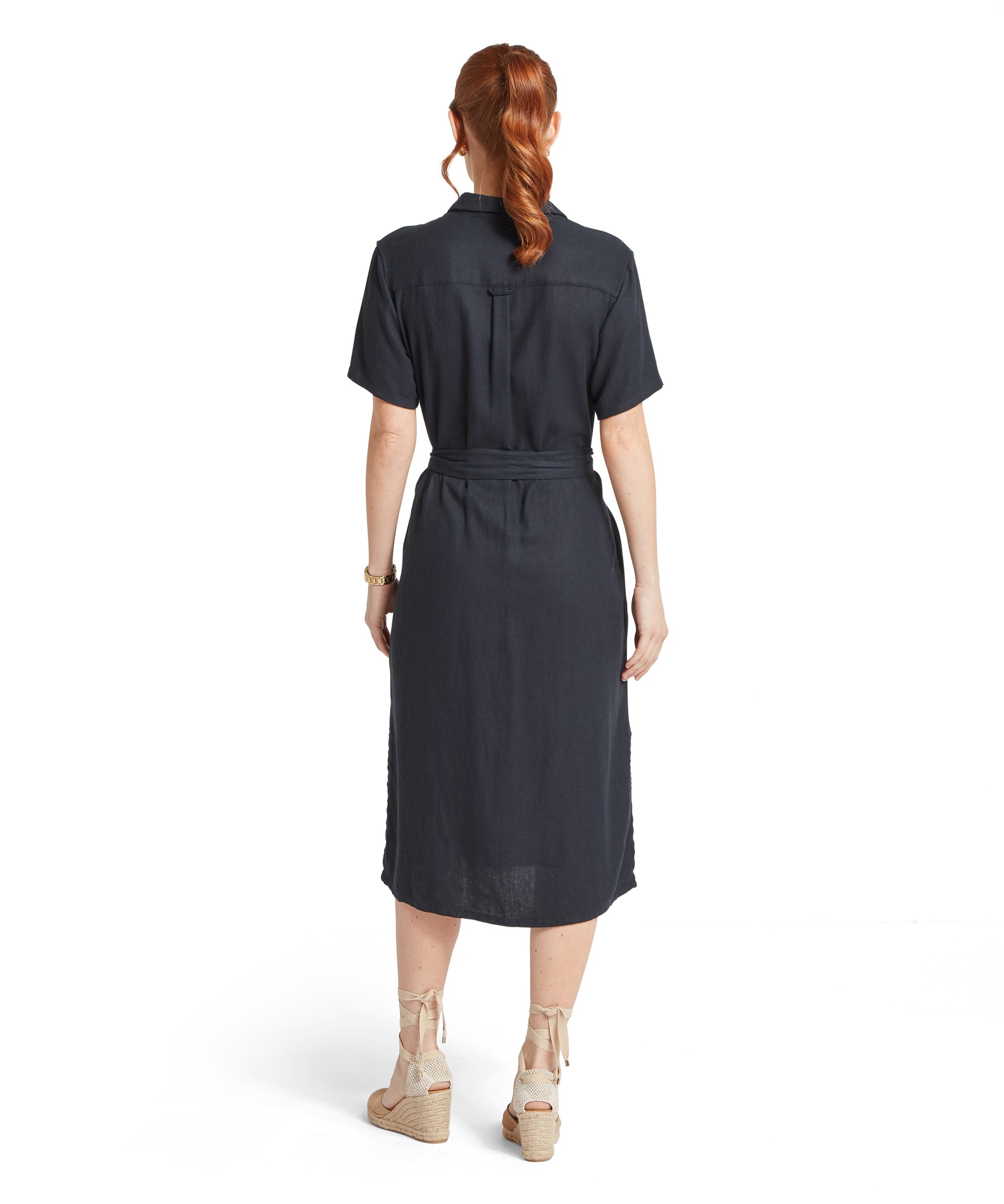 Back view of a woman standing against a plain white background wearing a navy linen short‑sleeve midi dress with a waist tie, paired with beige wedge sandals with ankle straps.