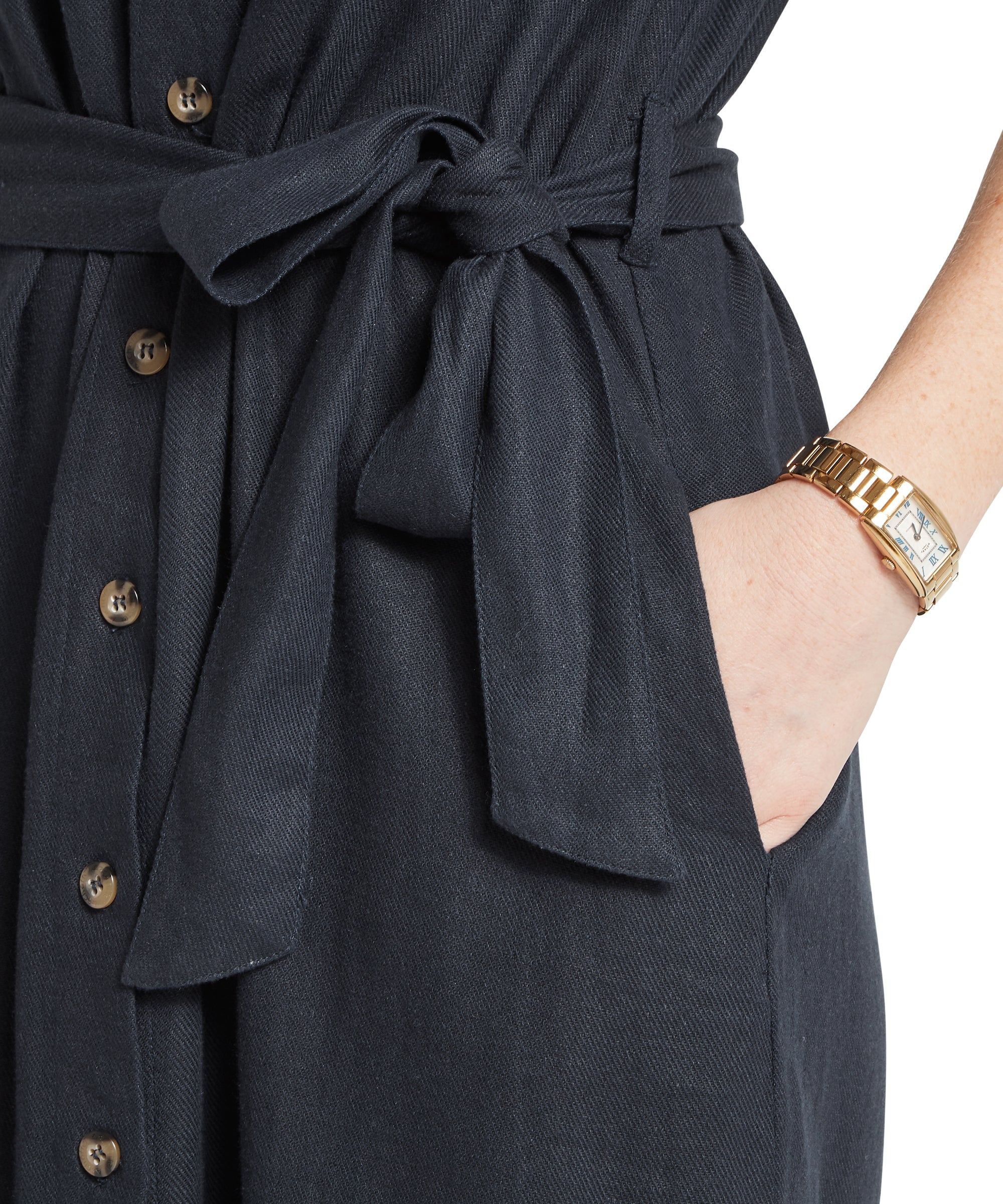 Close‑up of a woman wearing a navy textured button‑down dress with a tied waist and a hand in the pocket, shown with a gold rectangular‑face wristwatch.