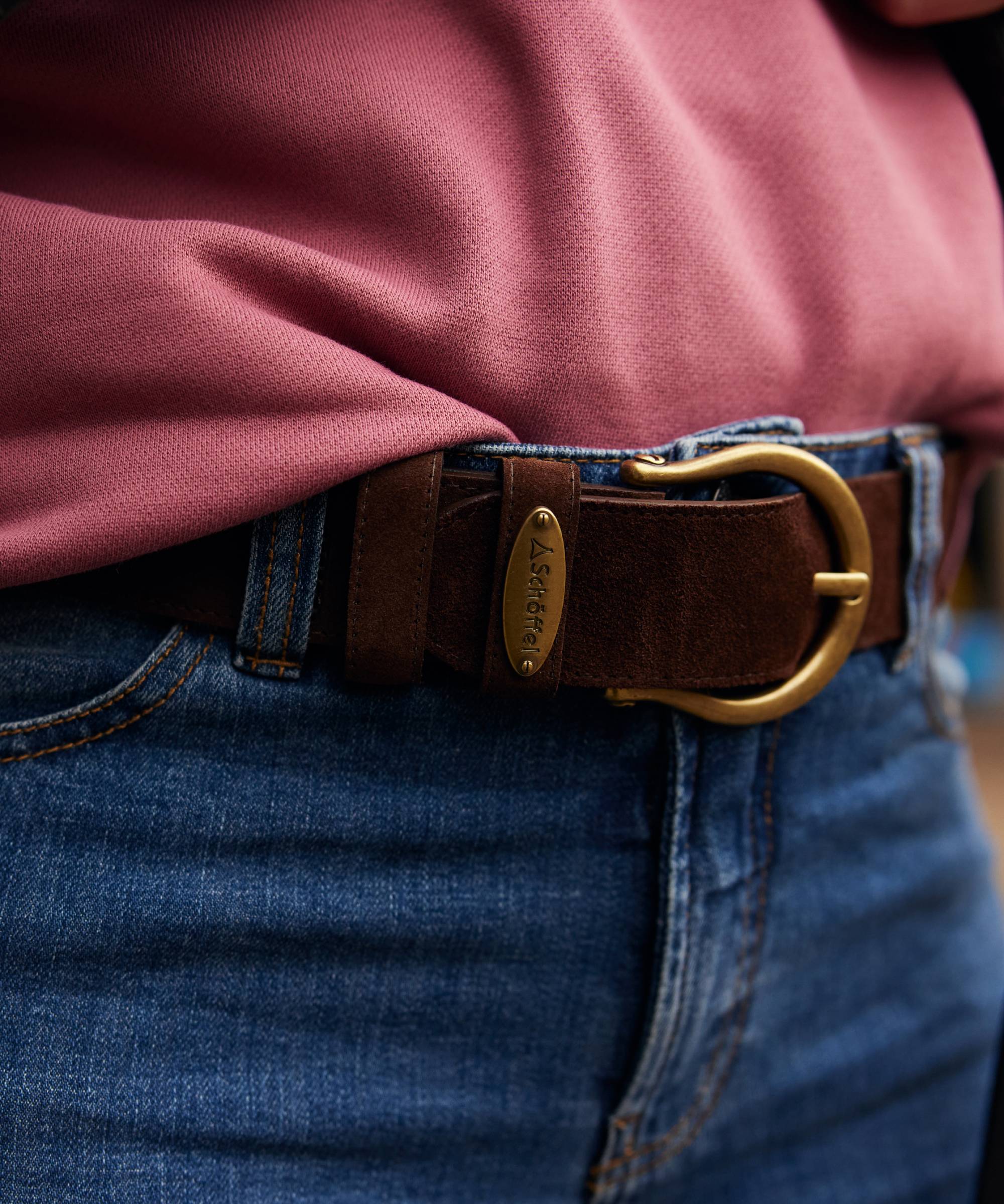 Close-up of a brown leather belt with gold buckle worn by a person in a pink shirt and blue jeans.
