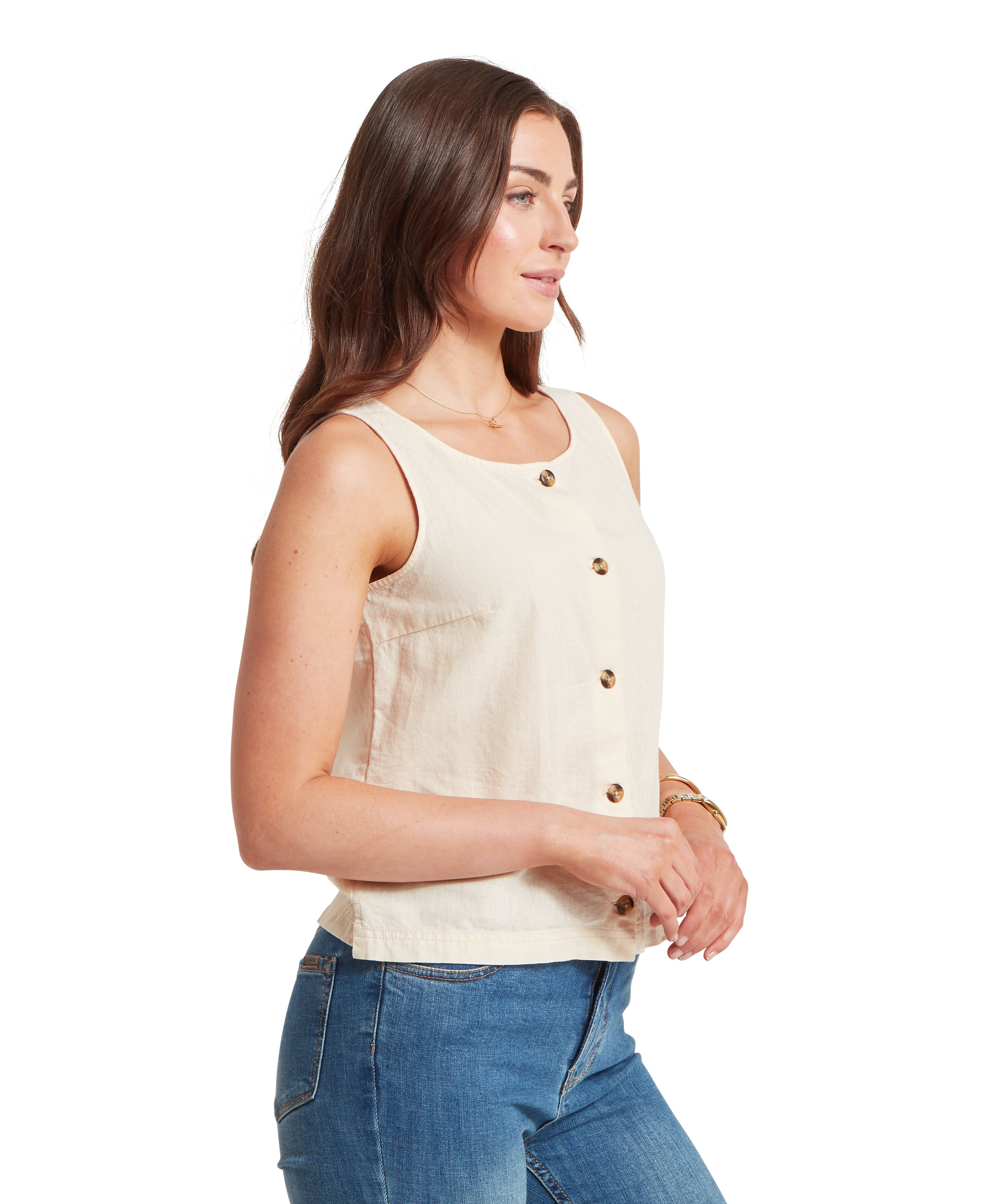 Woman standing in a side profile against a white background, wearing a cream-coloured sleeveless linen top. The top features a front placket with four small, dark tortoiseshell-style buttons and a straight hemline that sits at the hip. She is wearing blue denim jeans, and her long brown hair is styled over her shoulder. She is accessorised with a gold necklace and gold bracelets on her left wrist.