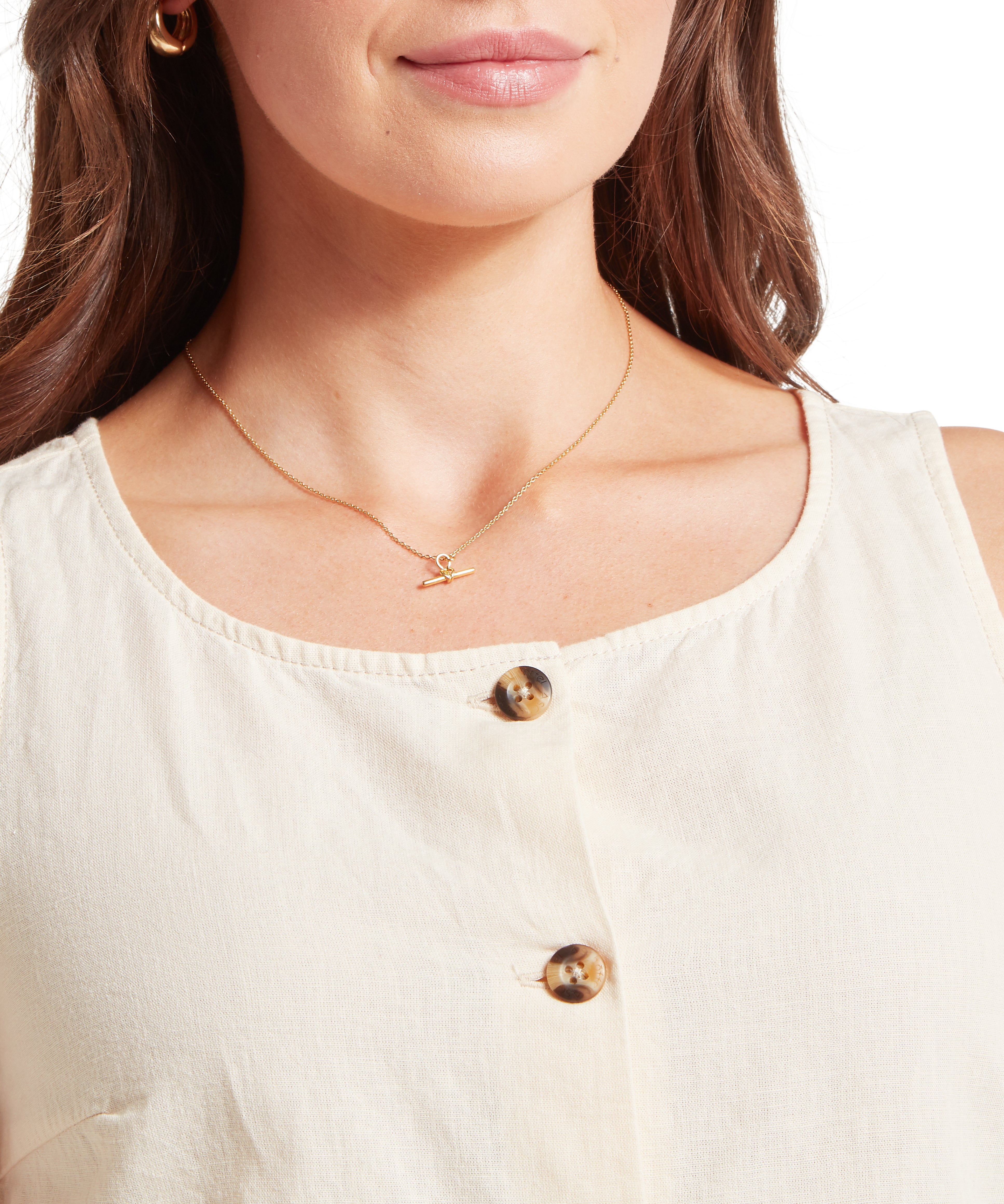 Woman's neckline is shown in a close-up, highlighting the round neck of a cream-coloured sleeveless linen top. The image displays a front placket with two visible dark tortoiseshell-style buttons and the textured, woven quality of the fabric. She is accessorised with a delicate gold chain necklace featuring a small bar pendant and small gold hoop earrings, with her long brown hair framing her face