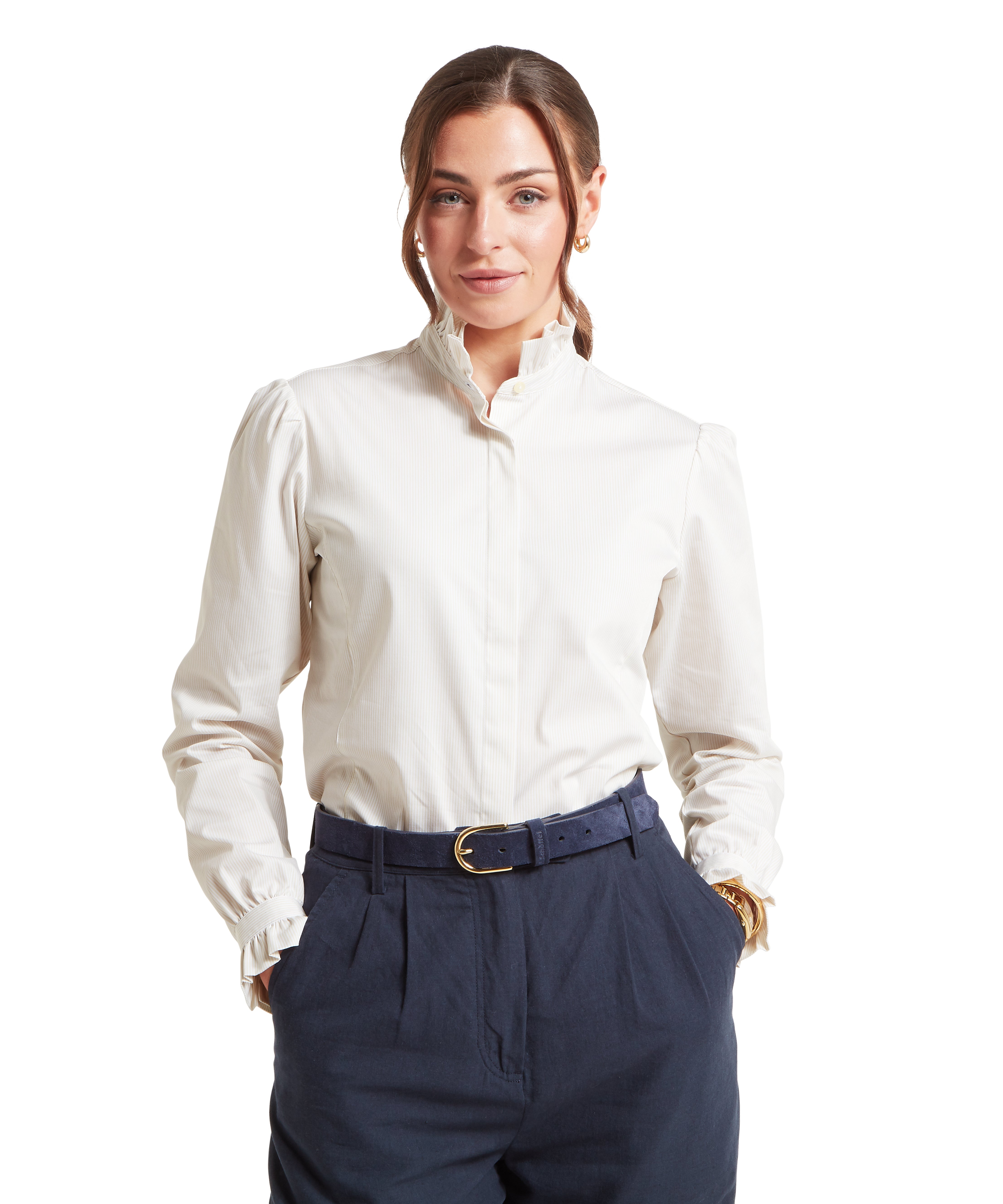 Woman wearing a beige blouse and navy trousers.  The woman is standing forwards with has both hands in her pockets and is in front of a white background.