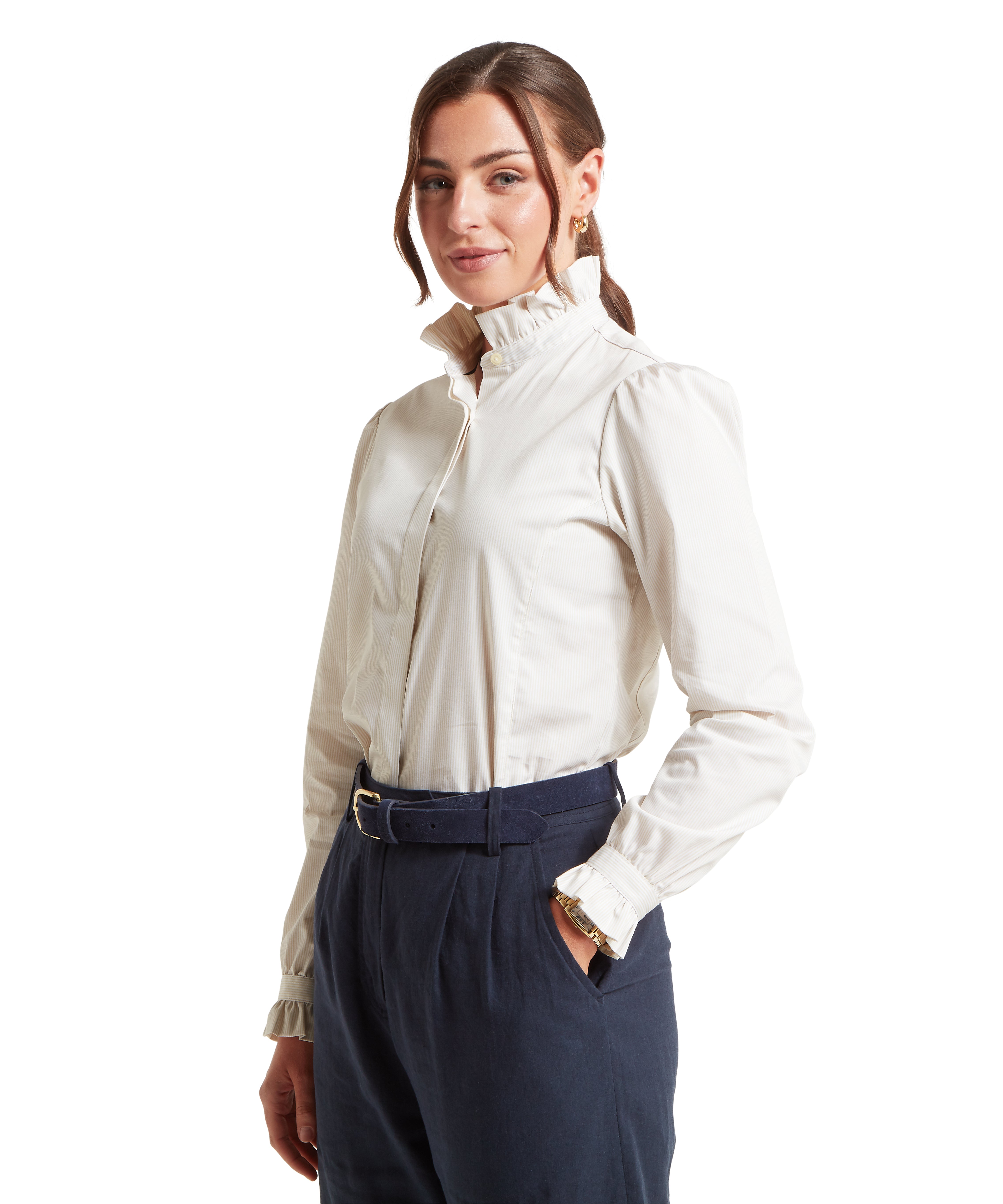 Woman standing in a three-quarter pose against a white background, wearing a white long-sleeved blouse with thin vertical stripes, featuring a high ruffled collar and matching ruffled cuffs. The blouse is tucked into high-waisted navy blue trousers, which are secured with a matching navy belt with a gold buckle. She has her left hand in her pocket, revealing a gold watch, and her hair is tied back with soft strands framing her face.
