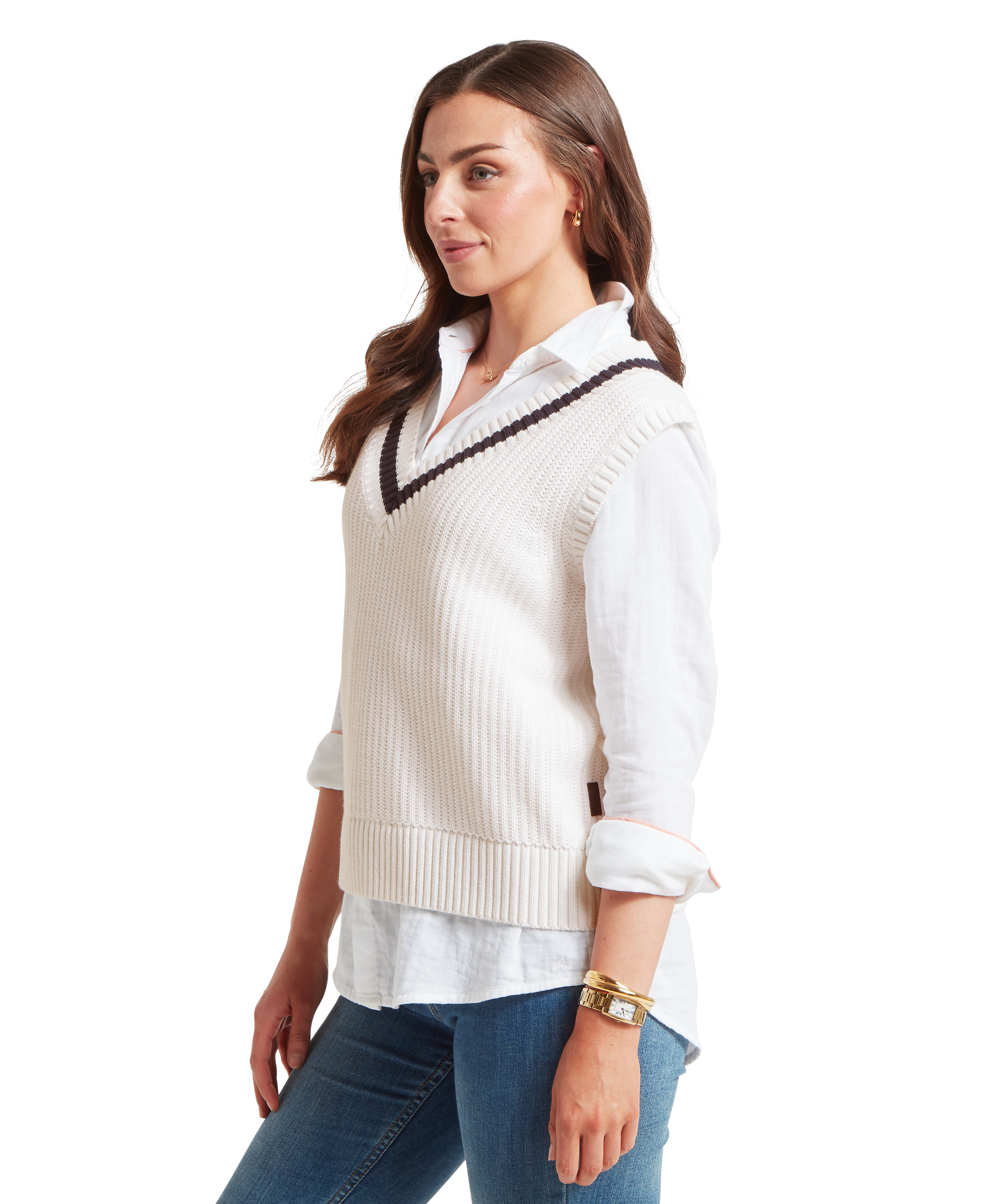 Woman facing slightly left, wearing a white collared shirt with rolled sleeves under a white knitted sleeveless V-neck vest with dark navy trim. Paired with blue jeans and a gold bracelet on left wrist. Plain white background highlights the layered styling and accessories.