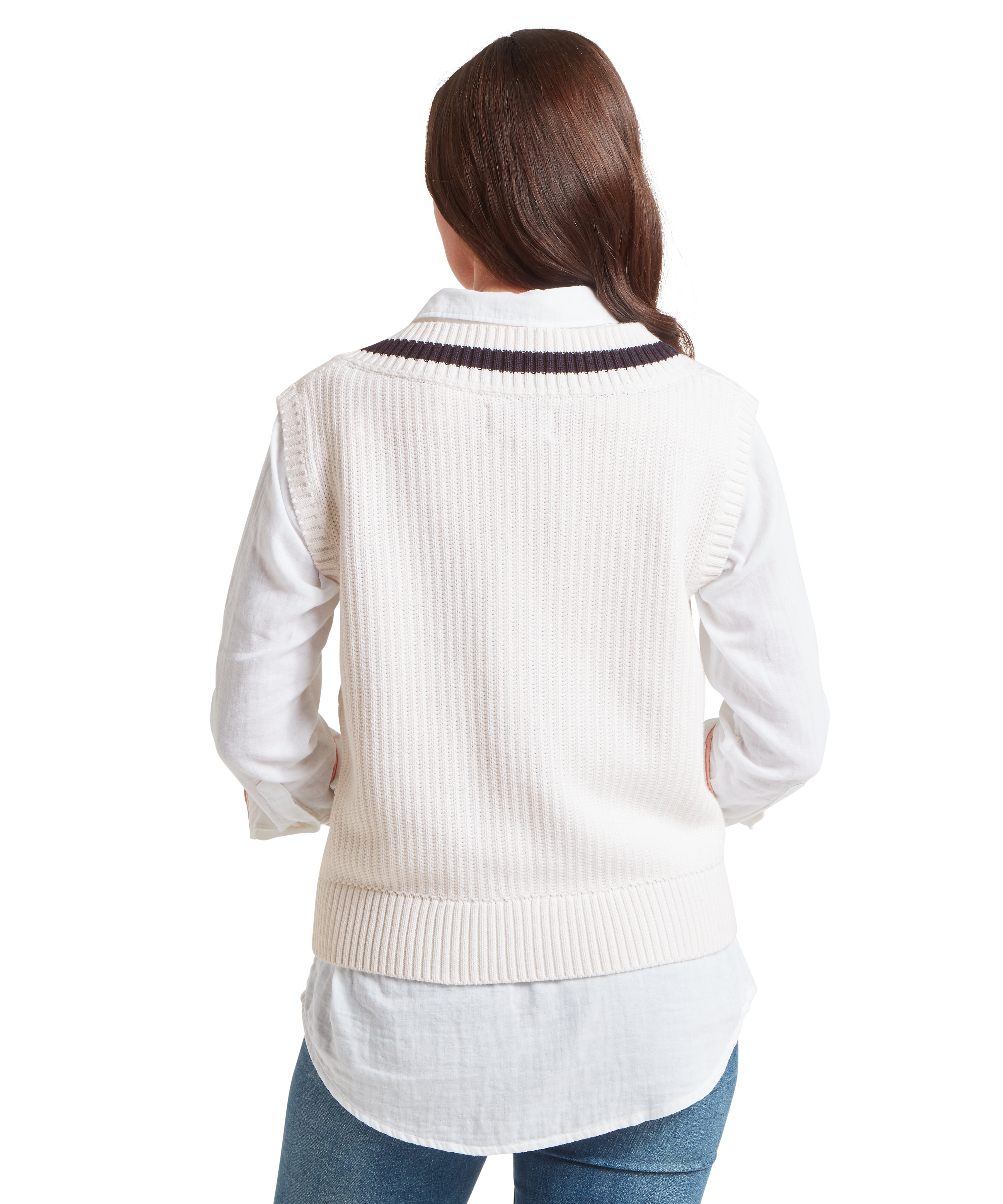 Rear view of a woman wearing a white sleeveless knitted jumper with ribbed hem and dark stripe detail near neckline, layered over a white long-sleeved button-up shirt. Paired with blue jeans. Plain white background highlights the textured knit and layered styling.