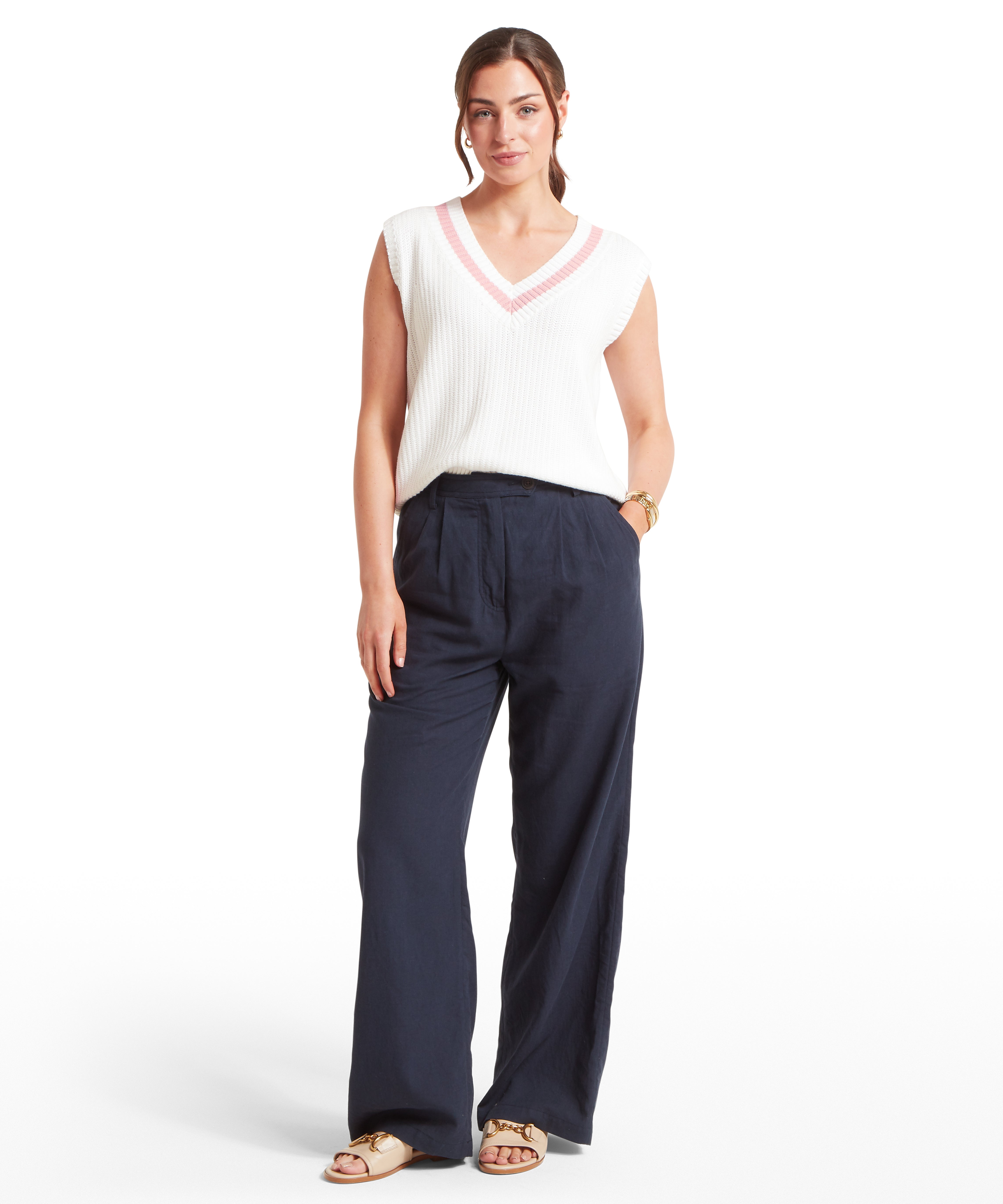 Woman wearing a white knitted vest with a V-neckline and  pink trim. Paired with high-waisted, wide-leg navy trousers, beige open-toe sandals, and a gold bracelet on the left wrist. Standing front-facing against a plain white background