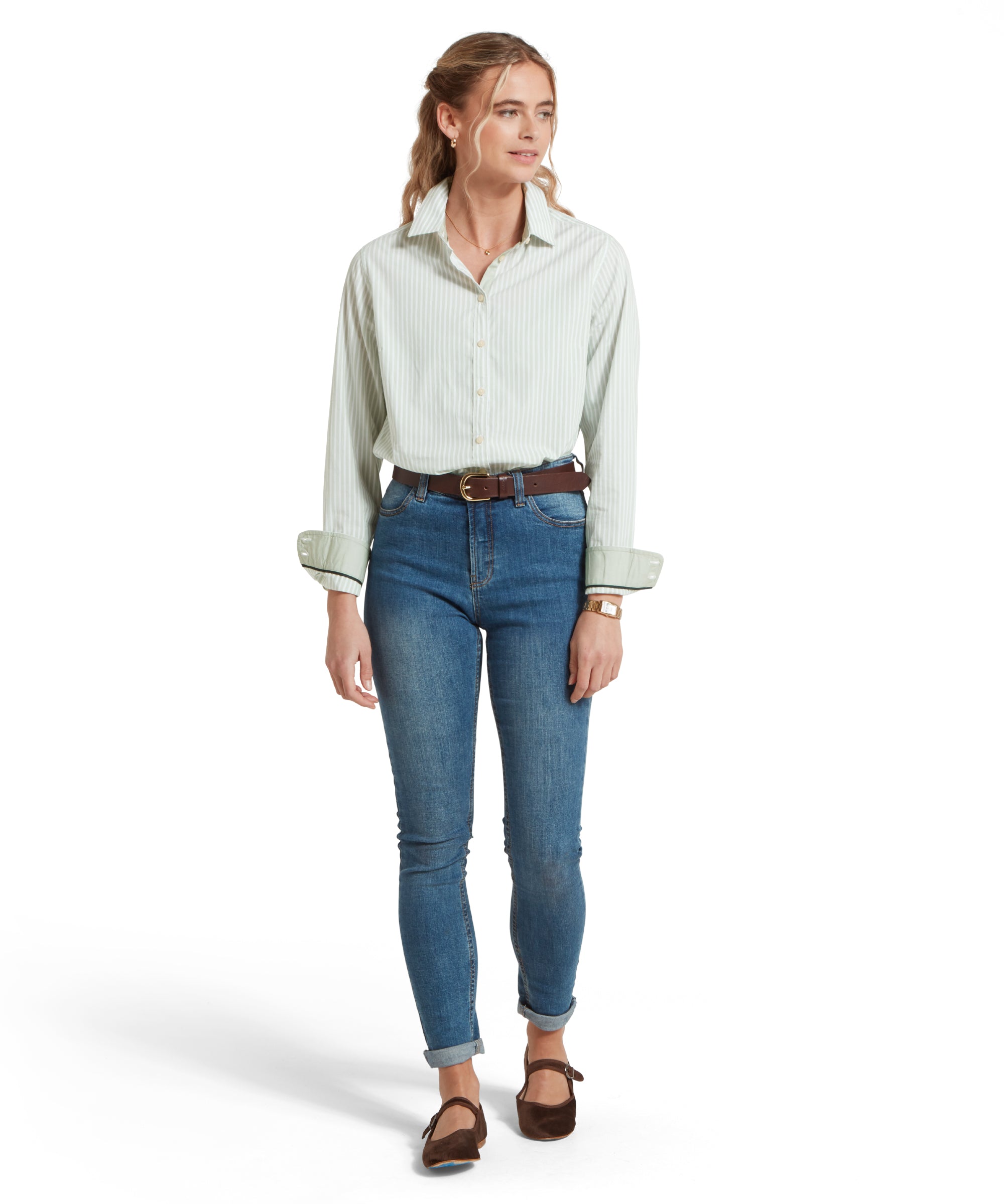 Full height of a female model wearing a green and white striped Schöffel shirt, paired with mid denim jeans, leather pumps and a tan leather belt. She is standing in a relaxed pose with her hands by his side.