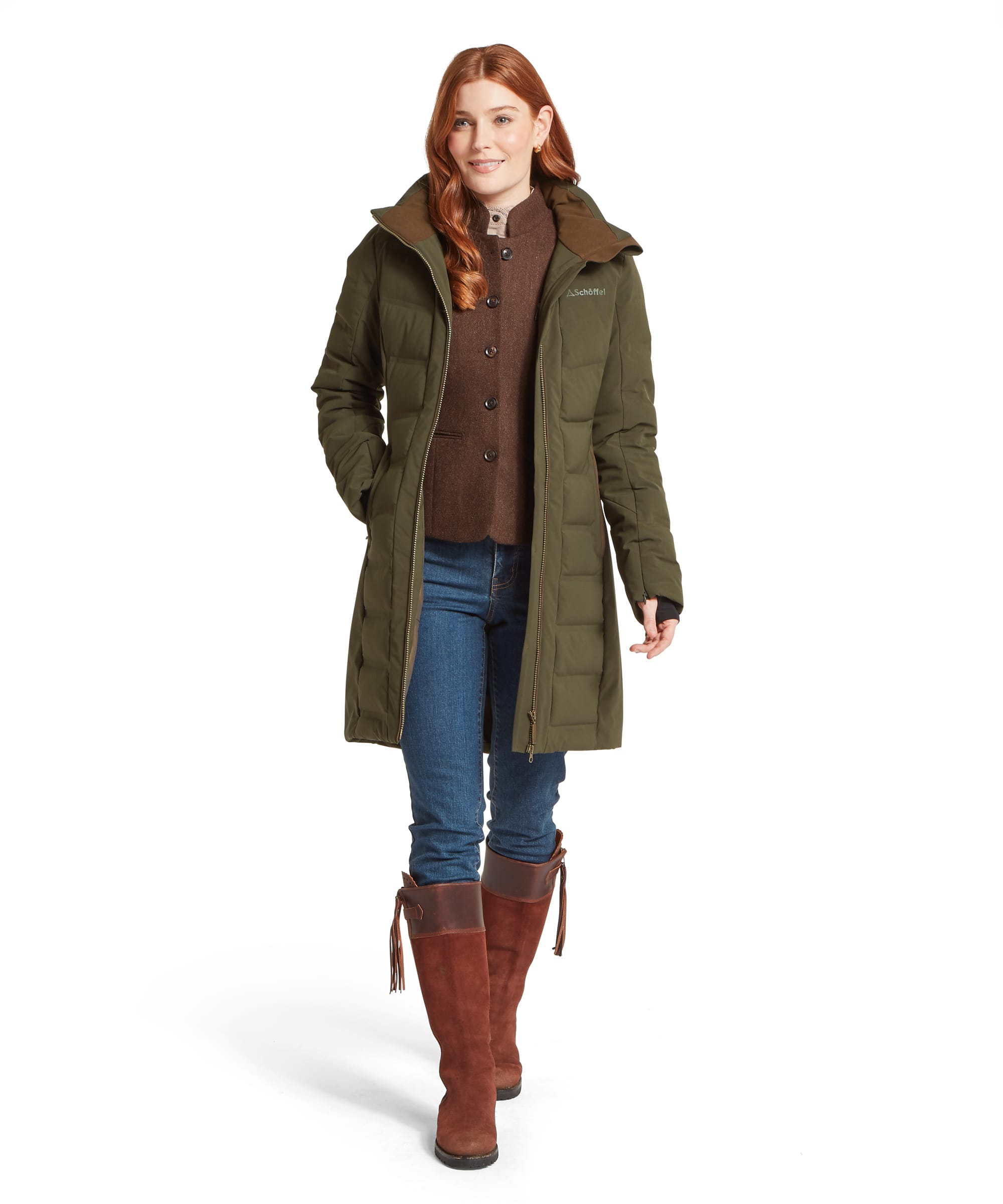 Full-length front view of a woman wearing the Schöffel Women's Hurlingham Waterproof Down Coat in Dark Olive, open to reveal layering over a brown tweed waistcoat and knitwear, styled with blue jeans and tan suede tassel riding boots