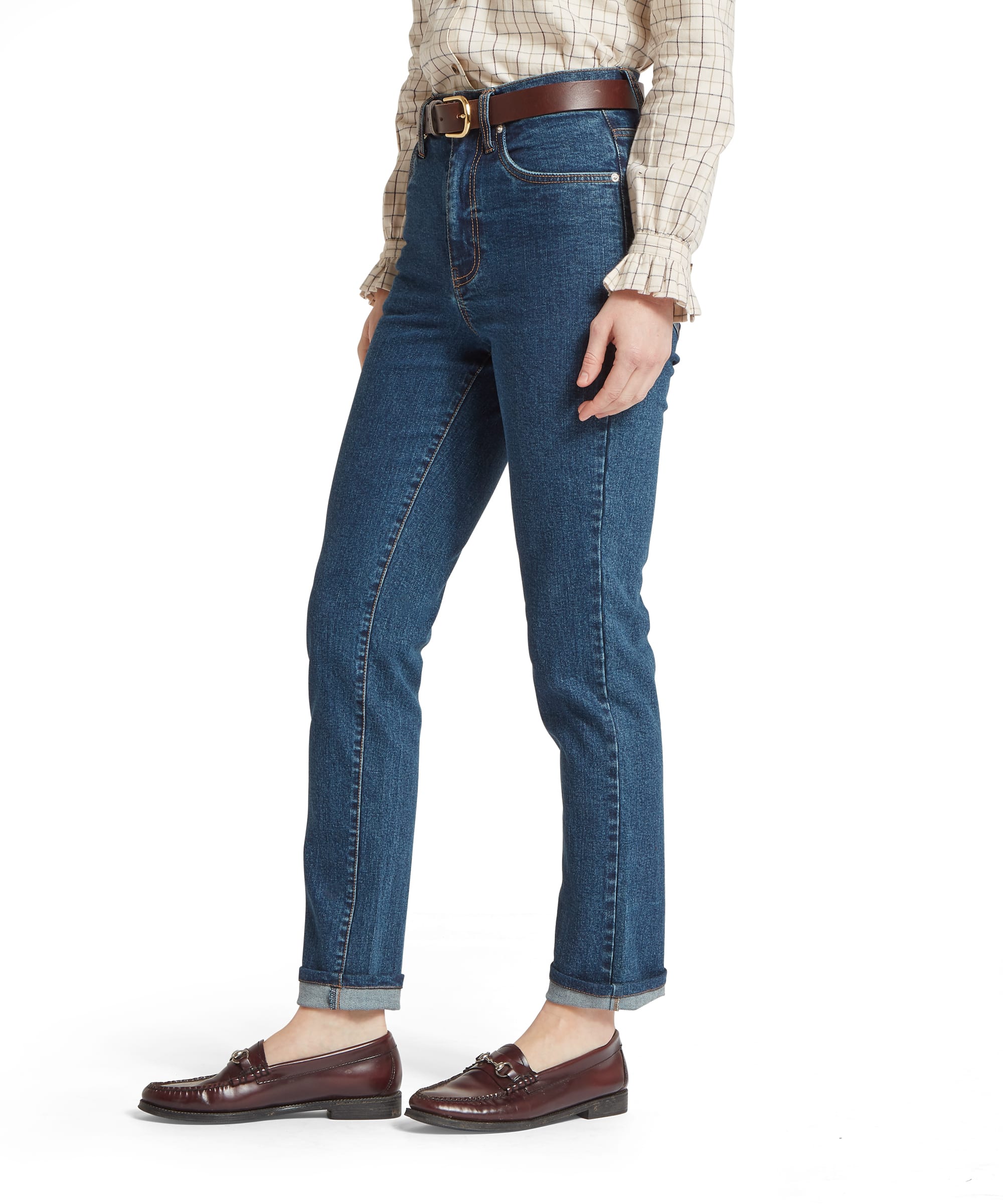 Schöffel Women's Ivy Jean in mid blue denim, side view showing high-rise waistband with brown leather belt, straight leg cut with turn-up hem, five-pocket styling with amber contrast stitching, worn with a cream windowpane-check shirt and dark horsebit loafers