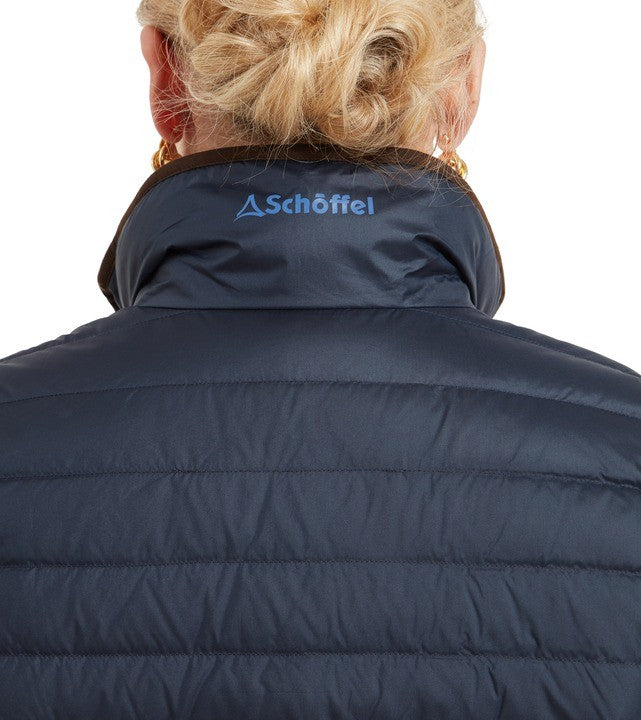 Rear view of a blonde woman with her hair up, wearing a navy blue quilted padded gilet by Schöffel. The Schöffel logo is printed in blue on the back of the collar. The gilet features horizontal quilted stitching across the back and a dark contrast inner collar lining.