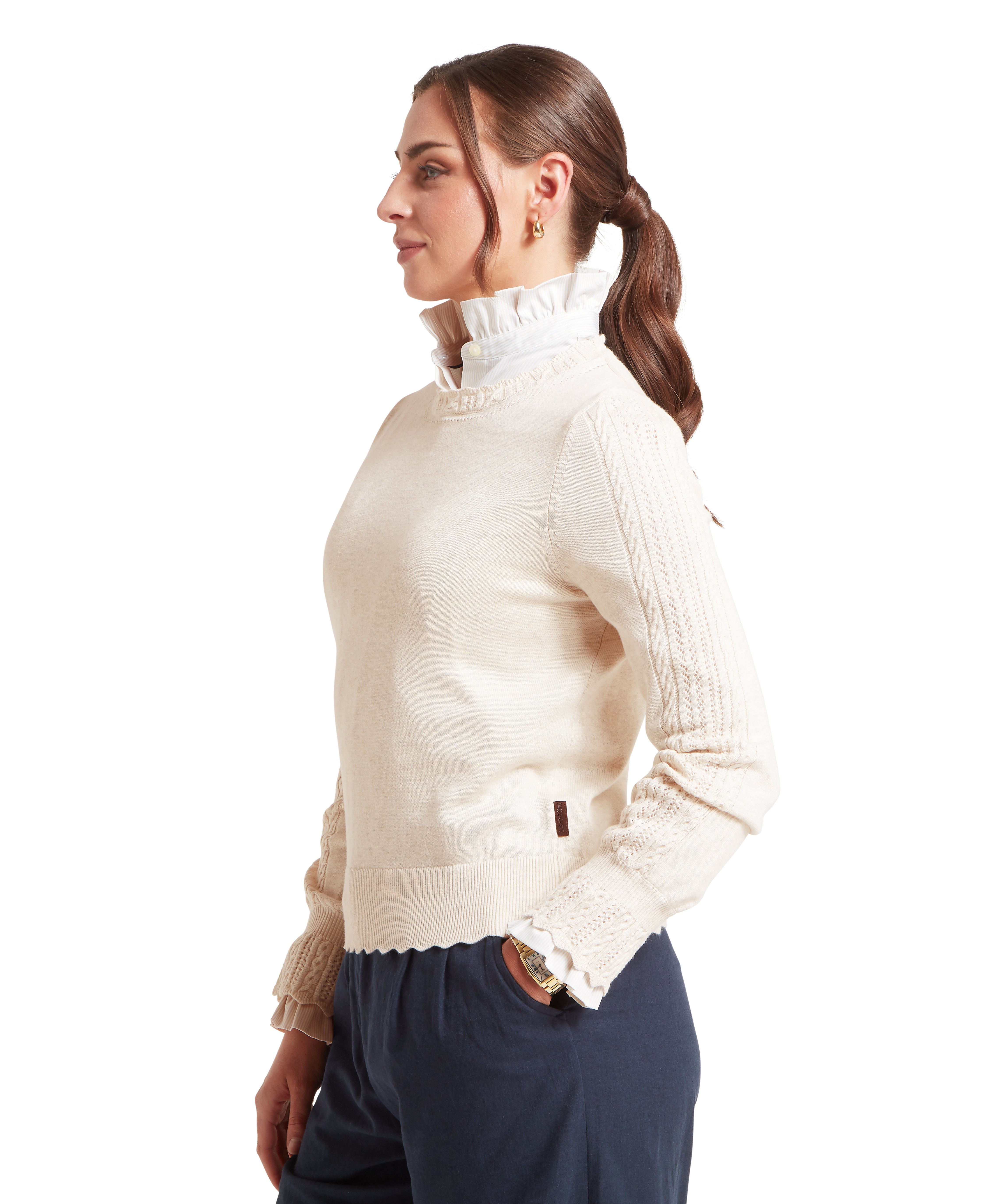 Profile view of a person wearing a cream-colored knitted jumper with textured sleeves paired with a  white high ruffled collar shirt and  dark blue trousers. The woman has long dark hair styled in a low ponytail and wears gold hoop earrings. One hand is placed in the pocket, and the background is plain white.