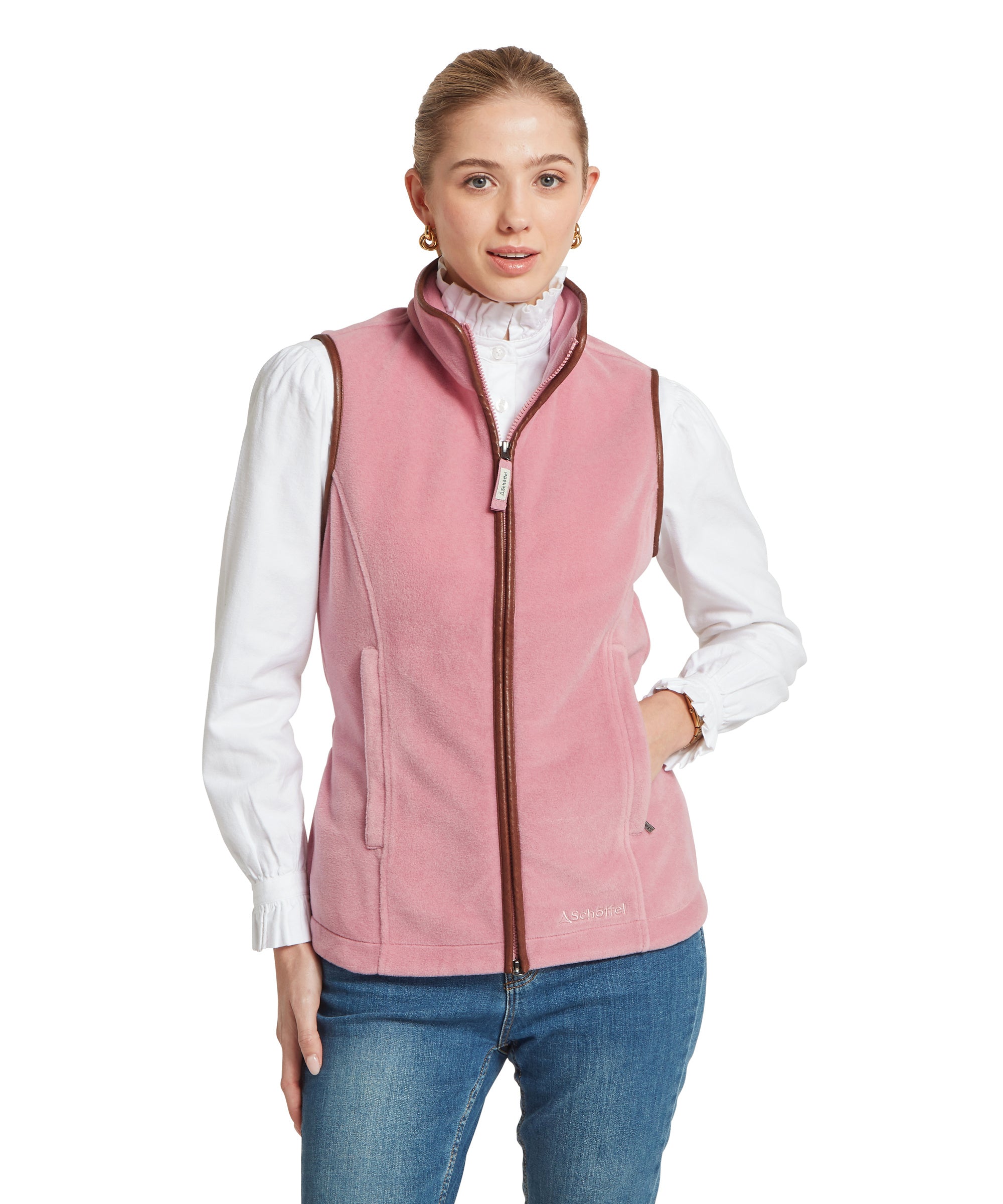 A woman wearing a pink sleeveless fleece gilet with brown trim and a zip-up high collar, styled over a white long-sleeved blouse and blue jeans, standing against a white background.