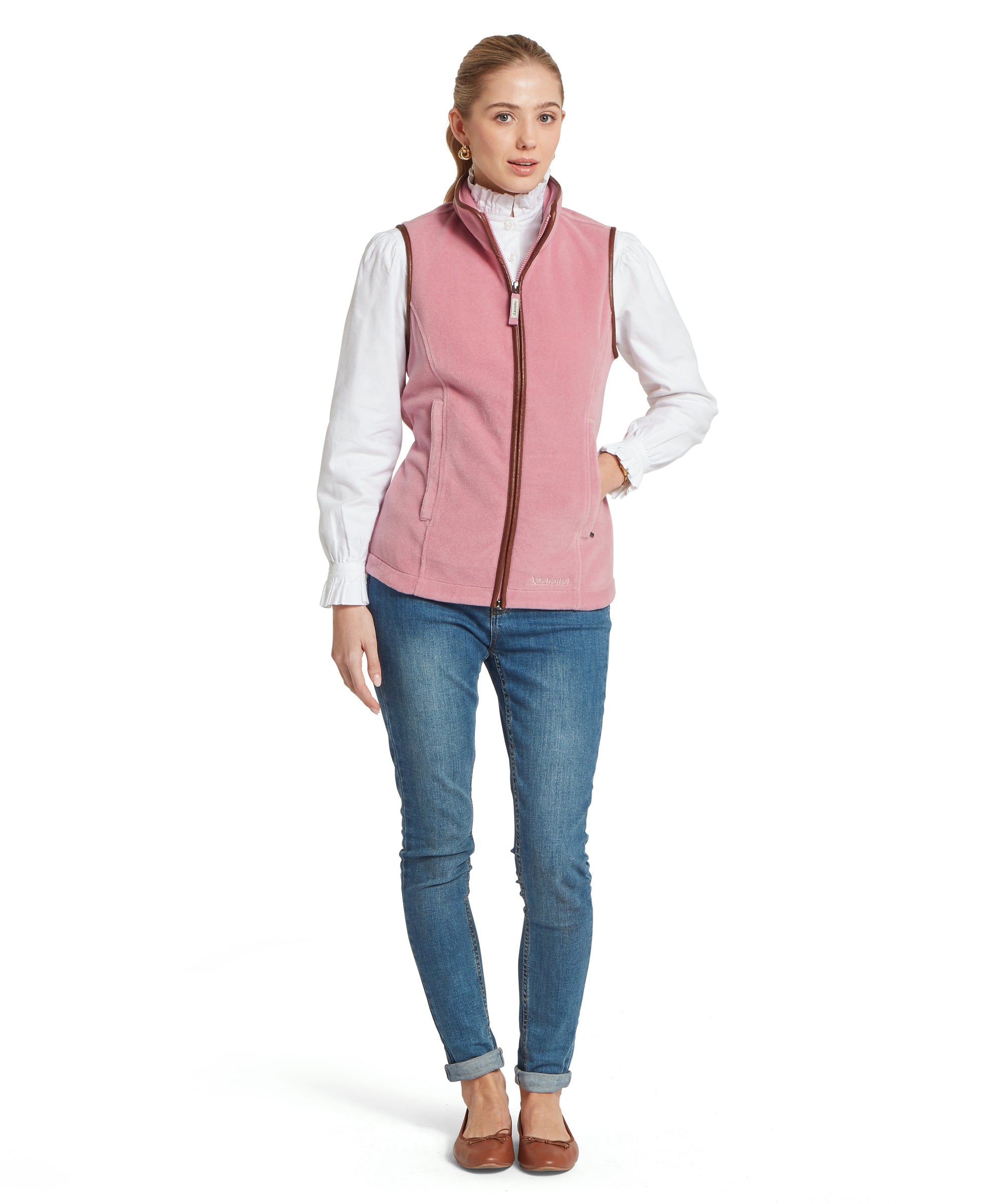 A woman wearing a light pink zip-up fleece vest with brown trim over a white long-sleeve blouse with puffed sleeves and a high collar. She pairs it with blue rolled-hem jeans and brown flat shoes. She stands facing forward against a plain background.