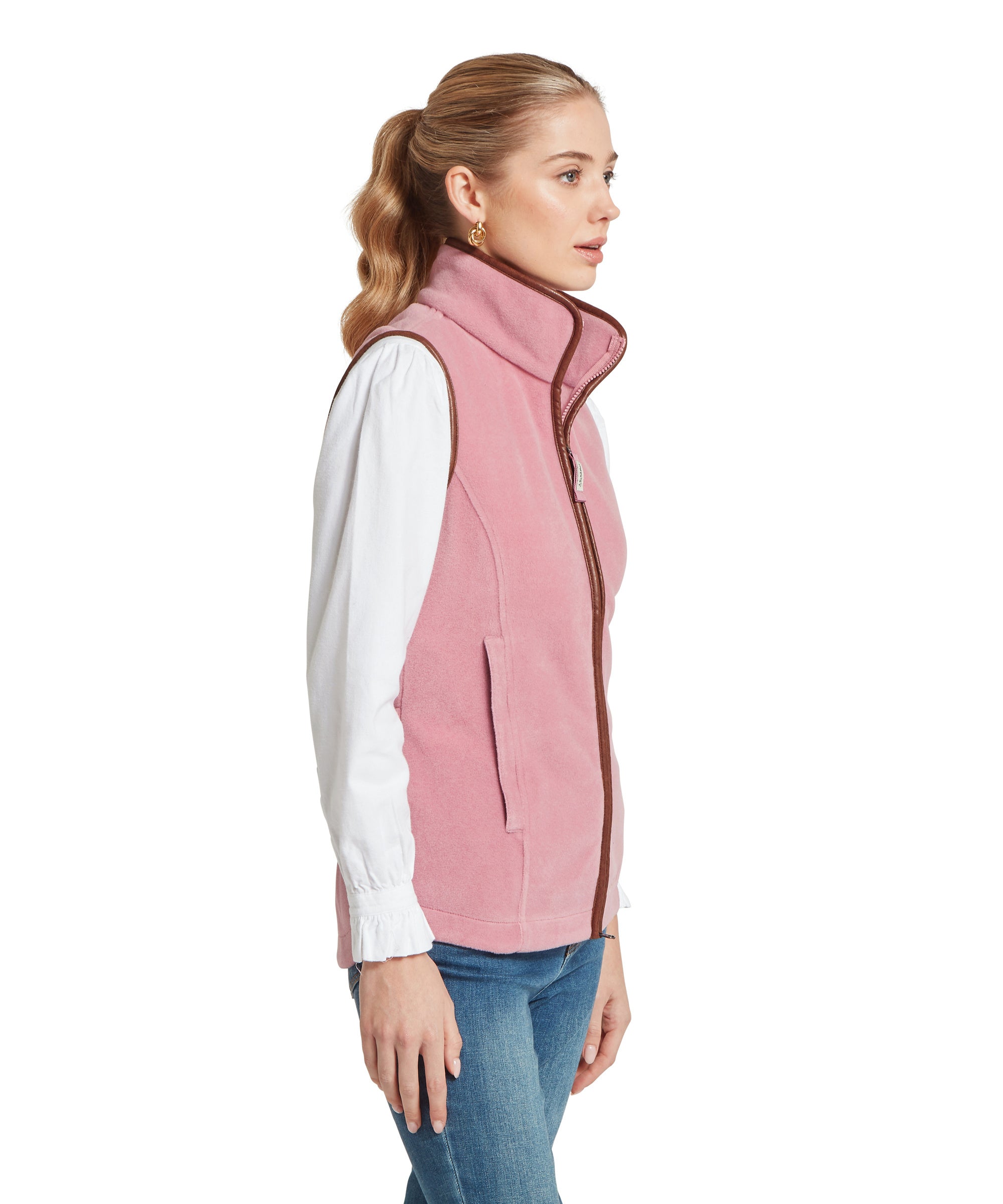A woman facing sideways wearing a light pink fleece vest with brown trim over a white long-sleeve blouse. The vest has a high collar and side pockets. She pairs it with blue jeans, and her hair is loosely waved and pulled back.