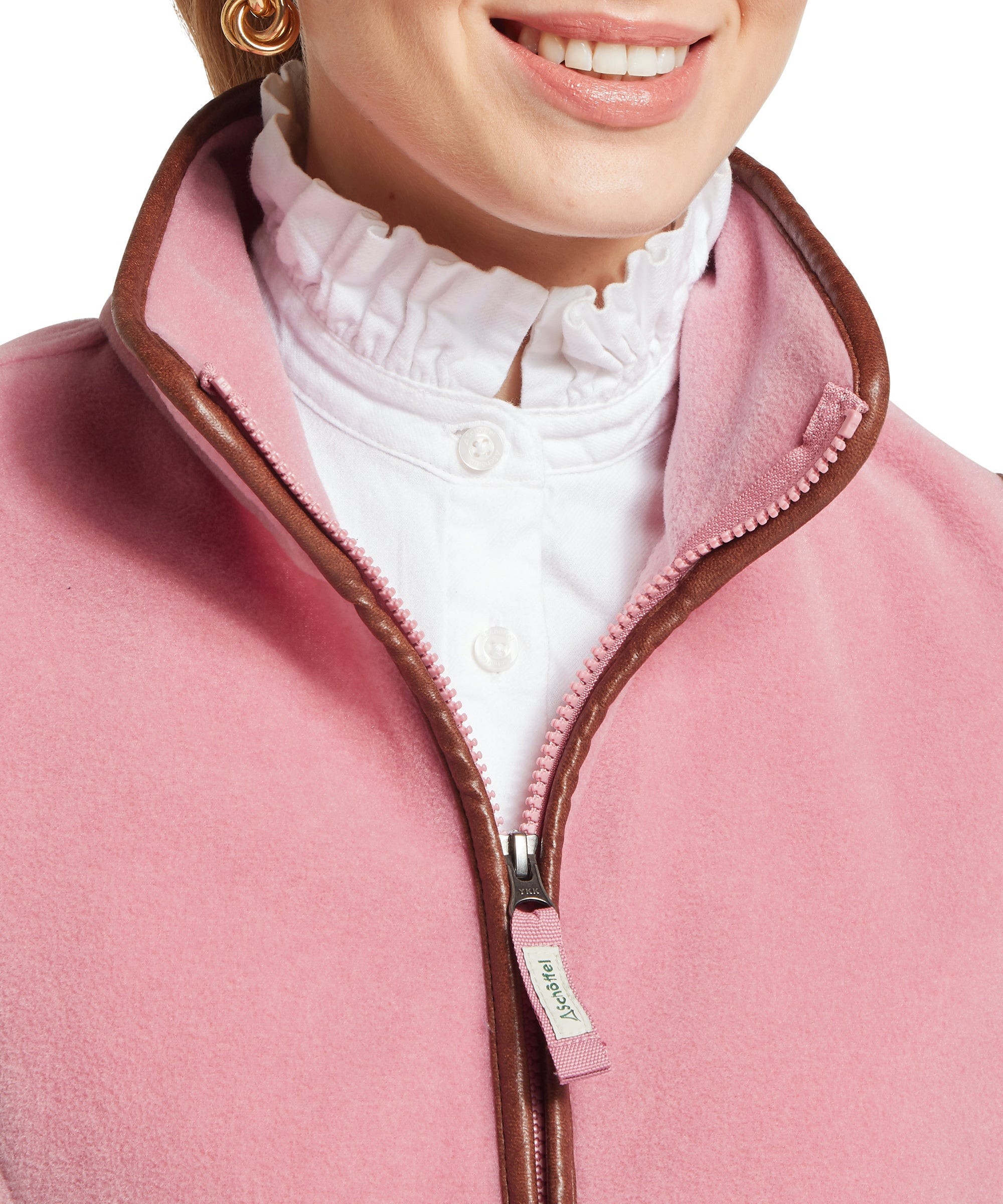 A close-up of a woman wearing a light pink fleece vest with brown trim, shown with the zip partially open to reveal a white blouse with a high ruffled collar. She wears gold hoop earrings.